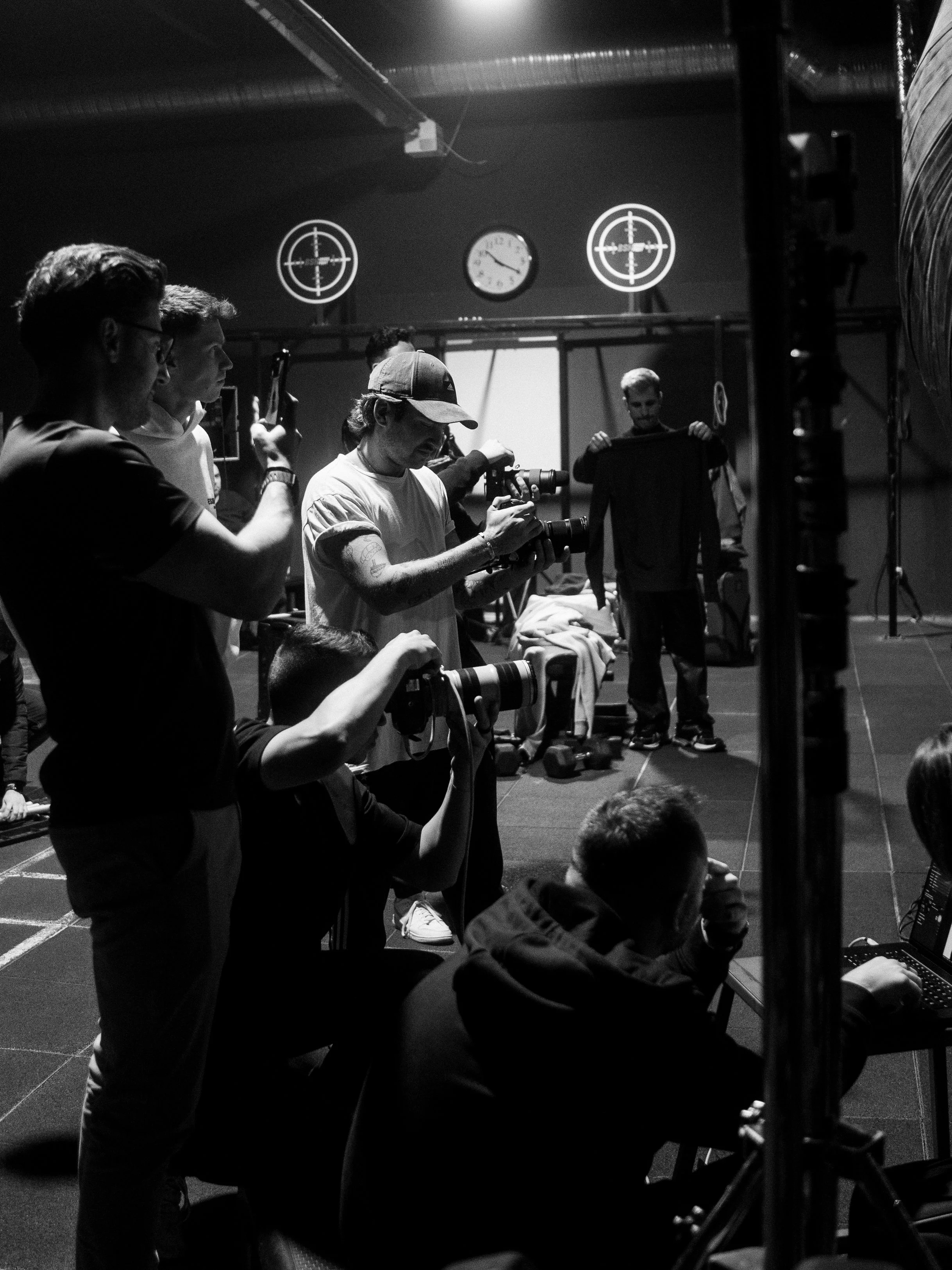 Black and white photo of a film set with crew members operating cameras and equipment, with two clocks on the wall showing different times.