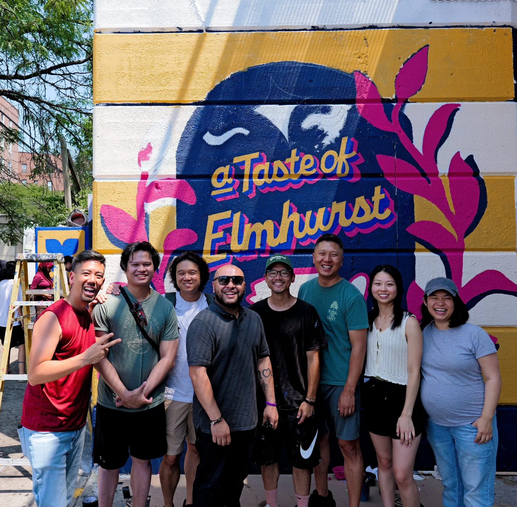 A group of nine diverse people smiling and posing in front of a colorful mural that reads 'A Taste of Elmhurst' with pink and purple floral designs.