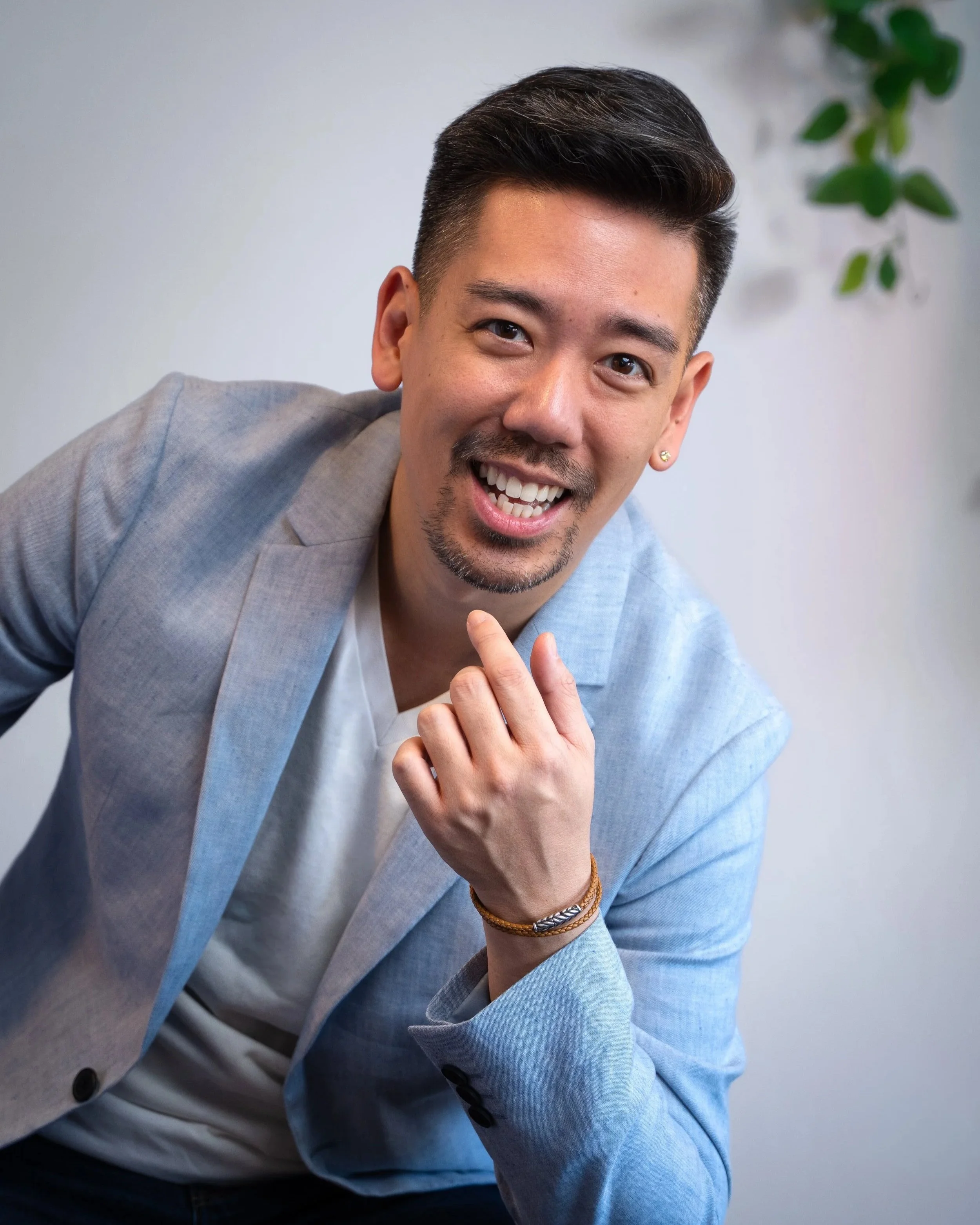 A smiling man with short dark hair, beard, and earrings, wearing a light gray blazer and white shirt, leaning forward and resting his chin on his hand, in front of a white wall with a green potted plant in the background.