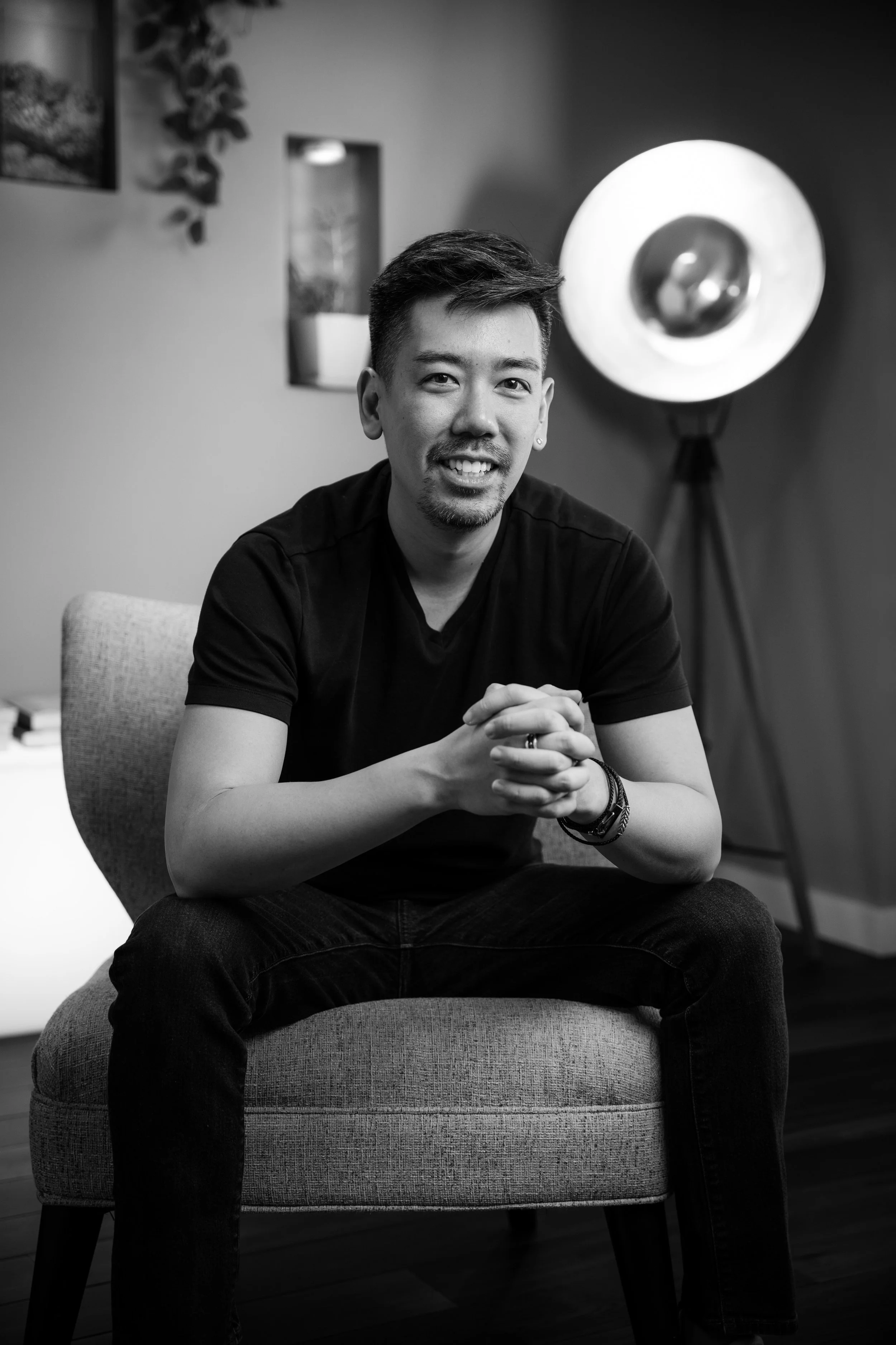 A man with short dark hair and a goatee, smiling with hands clasped, sitting on a cushioned chair in a modern interior, with a floor lamp and framed pictures in the background.