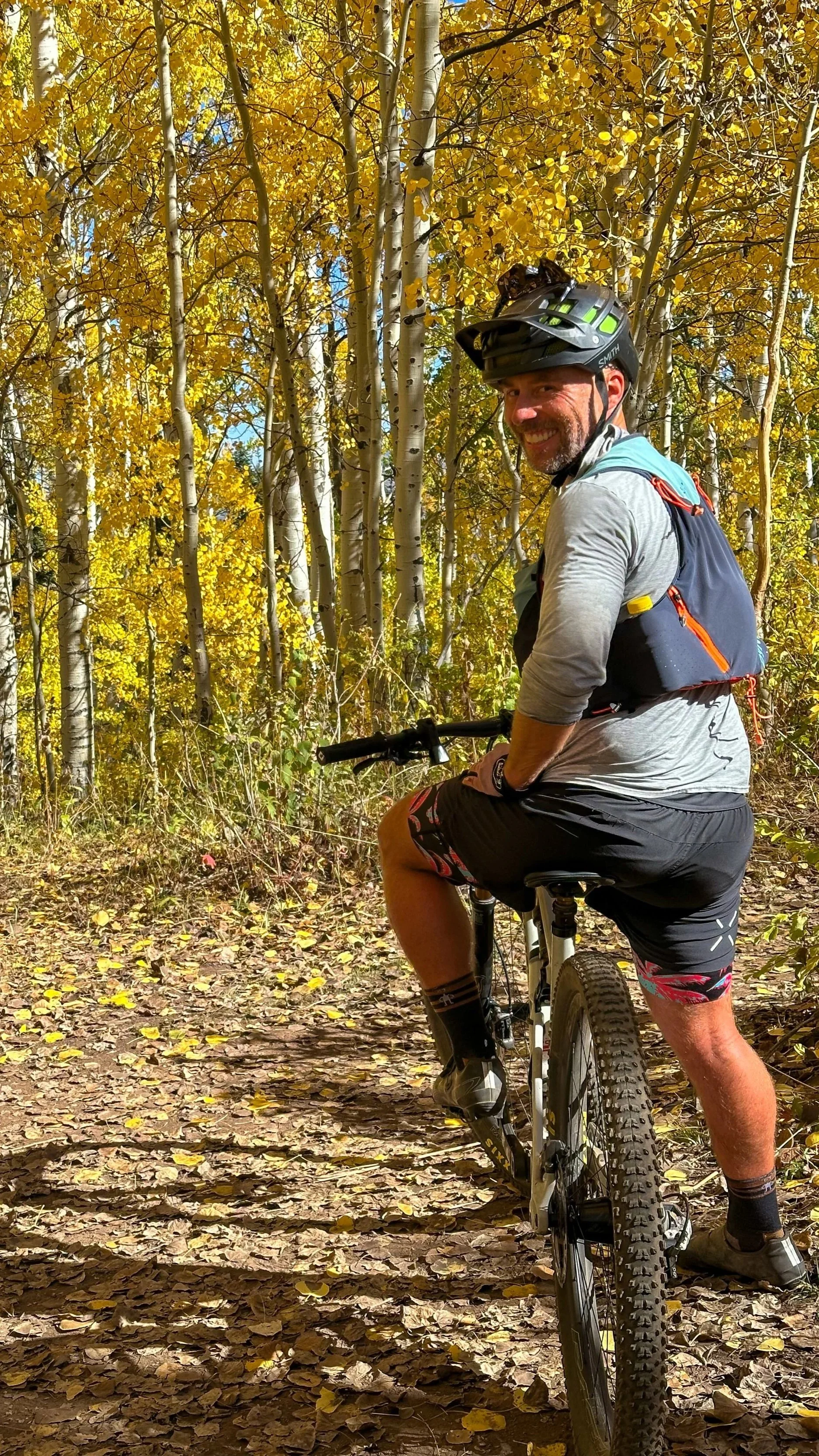 John Kucera riding a mountain bike on a leaf-covered trail in a forest during autumn, wearing a helmet, backpack, and athletic clothing, smiling at the camera.