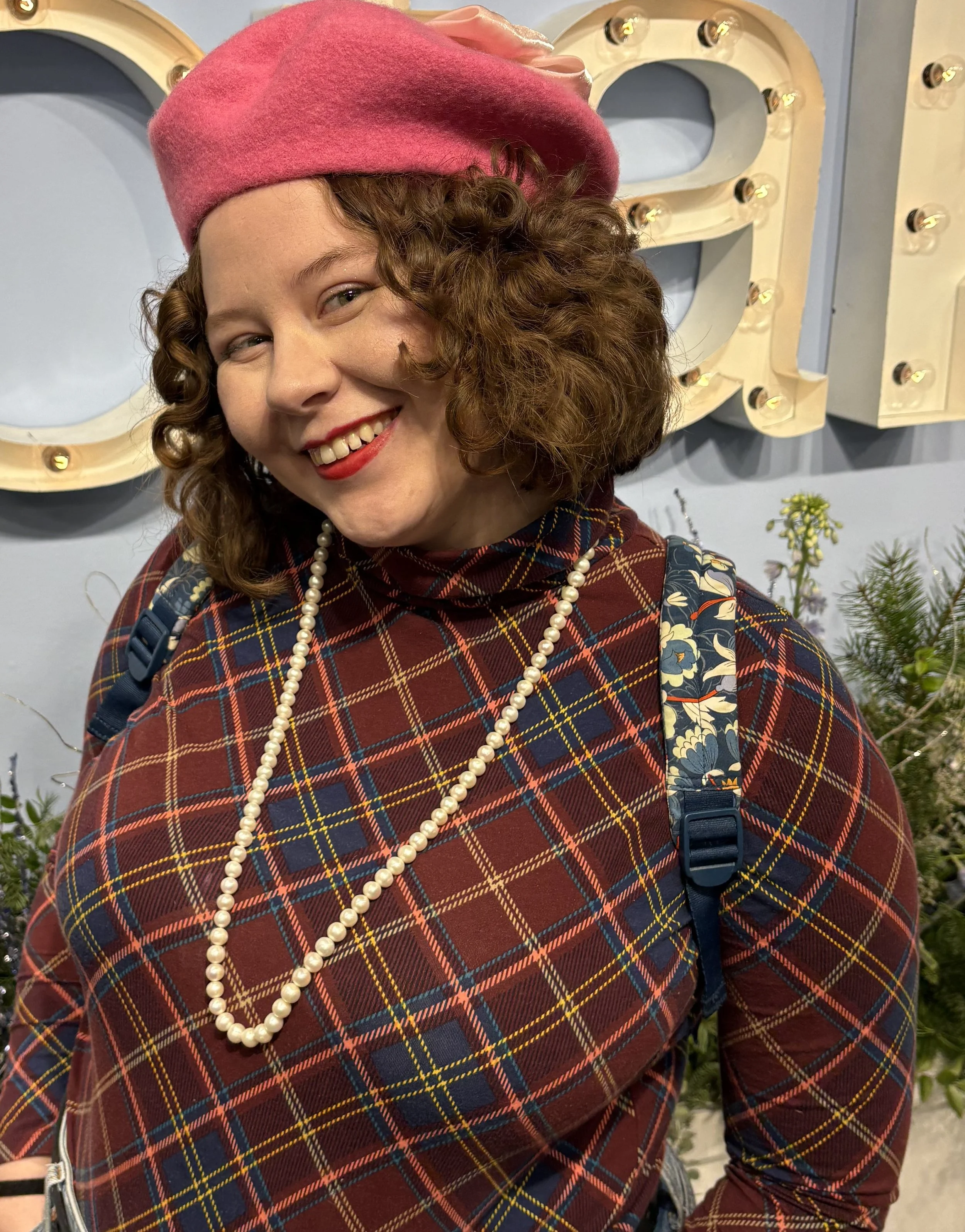 A woman with curly red hair wearing a pink beret, a plaid dress with red, navy, and yellow, a pearl necklace, and a blue floral backpack, smiling and standing in front of a light-colored wall with illuminated marquee-style letters.