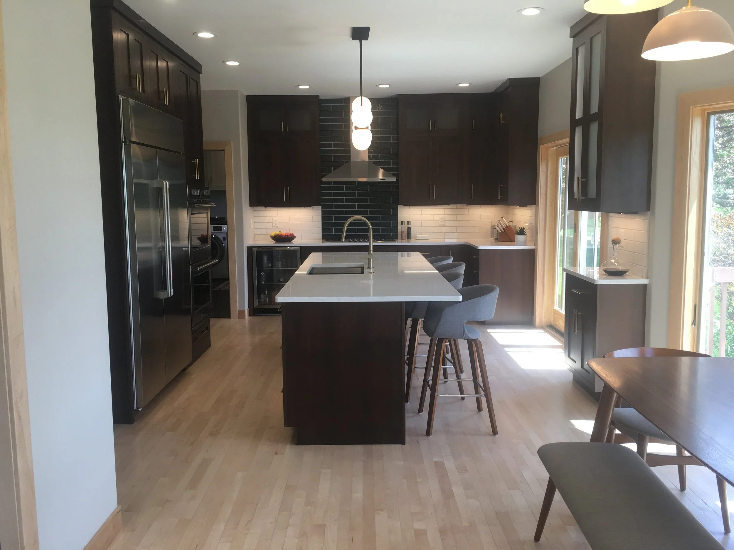 Modern kitchen with dark wooden cabinets, white island with bar stools, stainless steel appliances, black tiled backsplash, frosted glass light fixtures, wooden floor, large windows, and dining table.