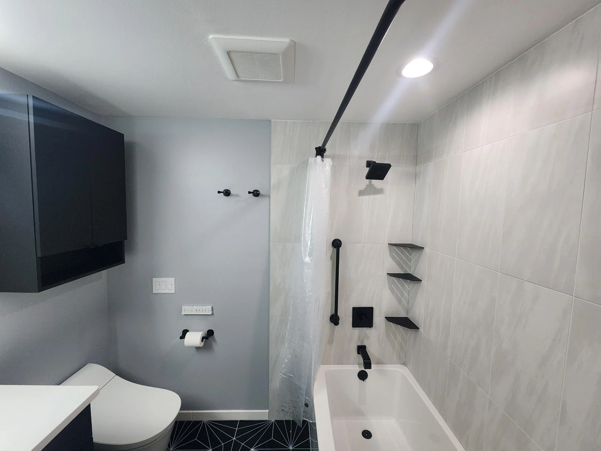 Modern bathroom with light-colored tiled walls, black fixtures including a showerhead, grab bar, and shelves, partial view of a toilet, black cabinet, and geometric black and white floor tiles.