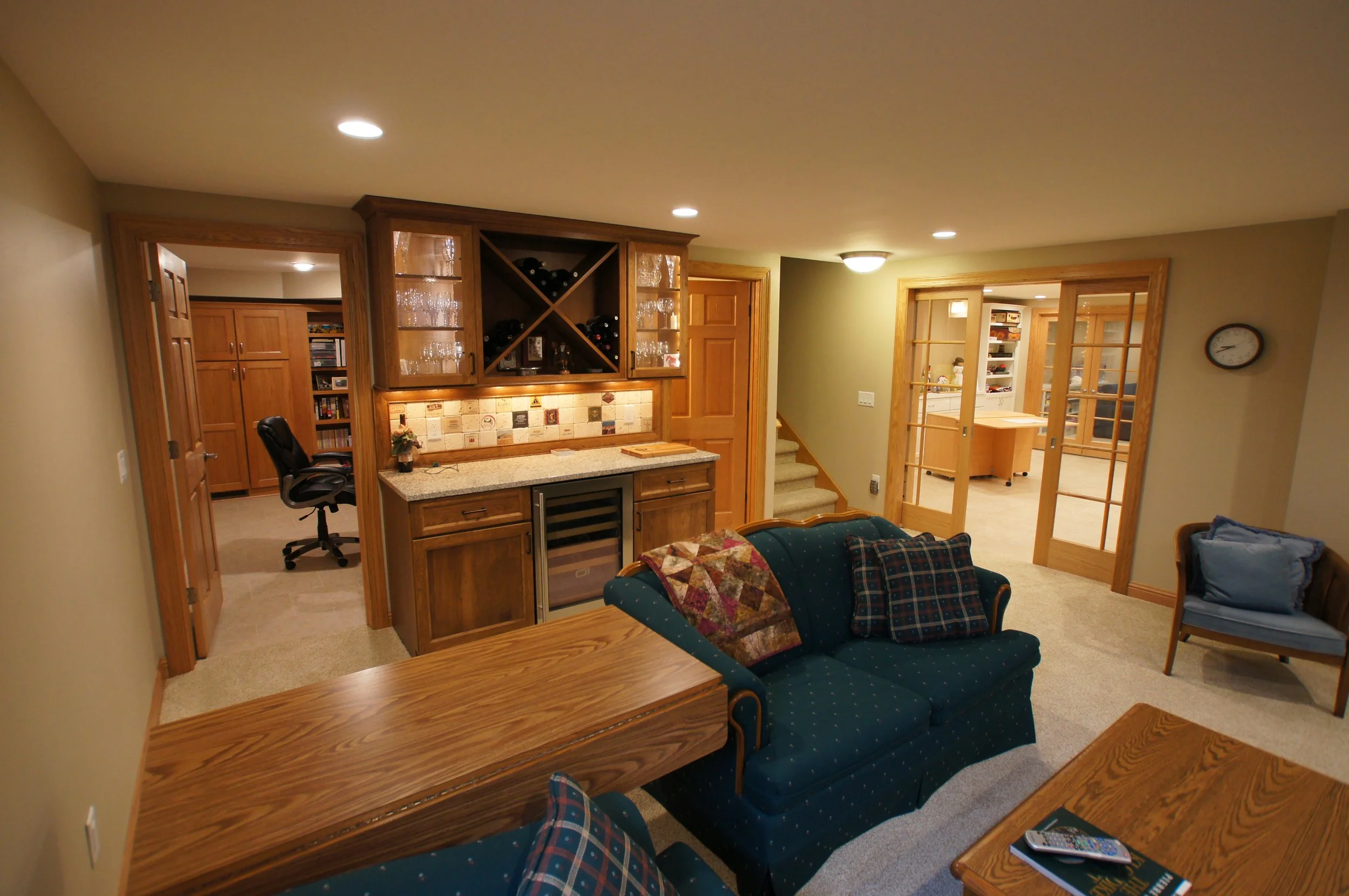 Living room area with blue upholstered sofa, armchair, wooden coffee table, and built-in wooden bar with shelves and glassware. There are sliding glass doors leading to a kitchen or dining area, a staircase, and a clock on the wall.