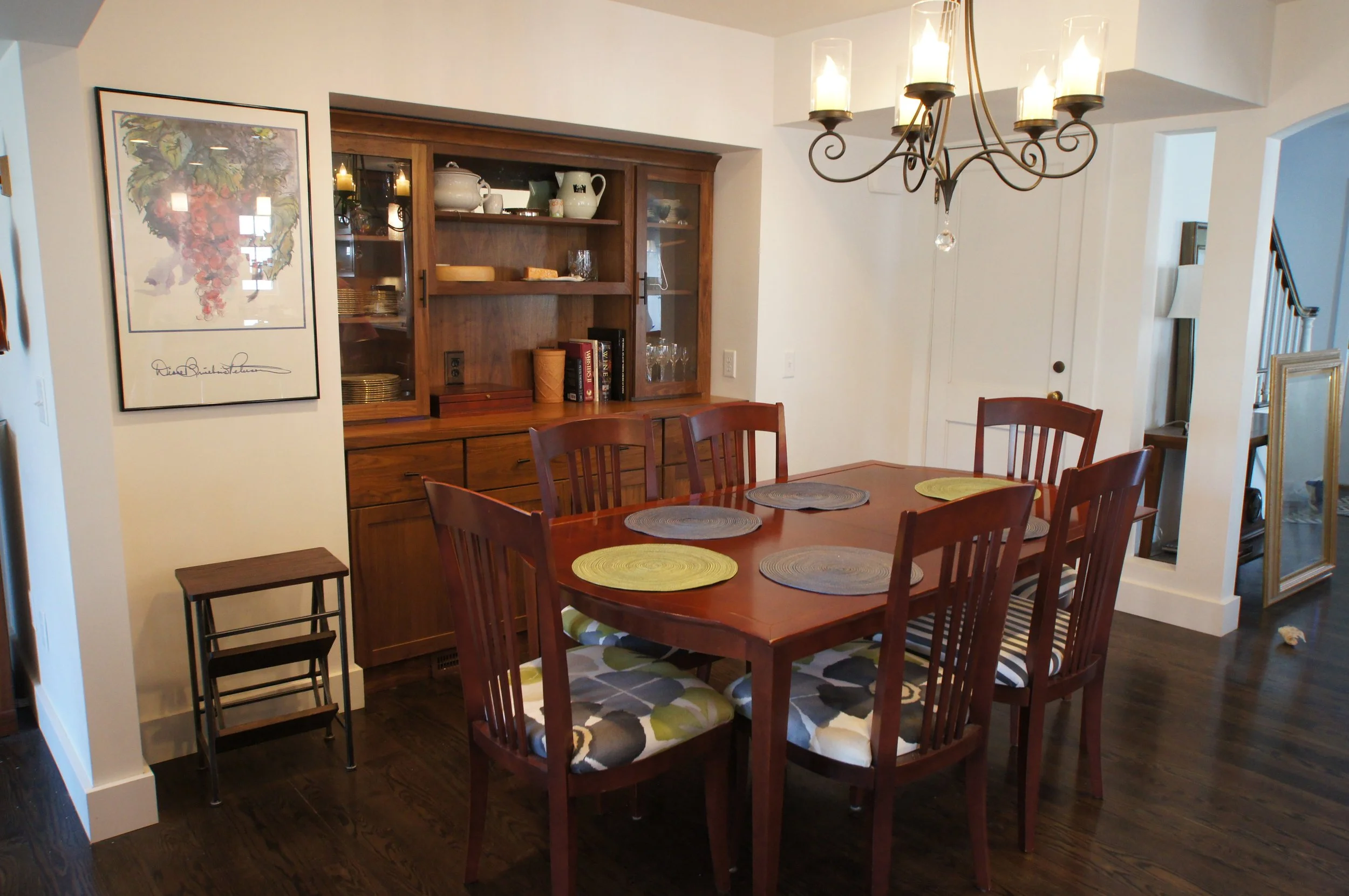A dining room with a wooden table and six chairs with patterned cushions. A chandelier hanging above the table, and a wooden china cabinet with glass doors and shelves behind it. There are placemats on the table, and a framed artwork on the wall.
