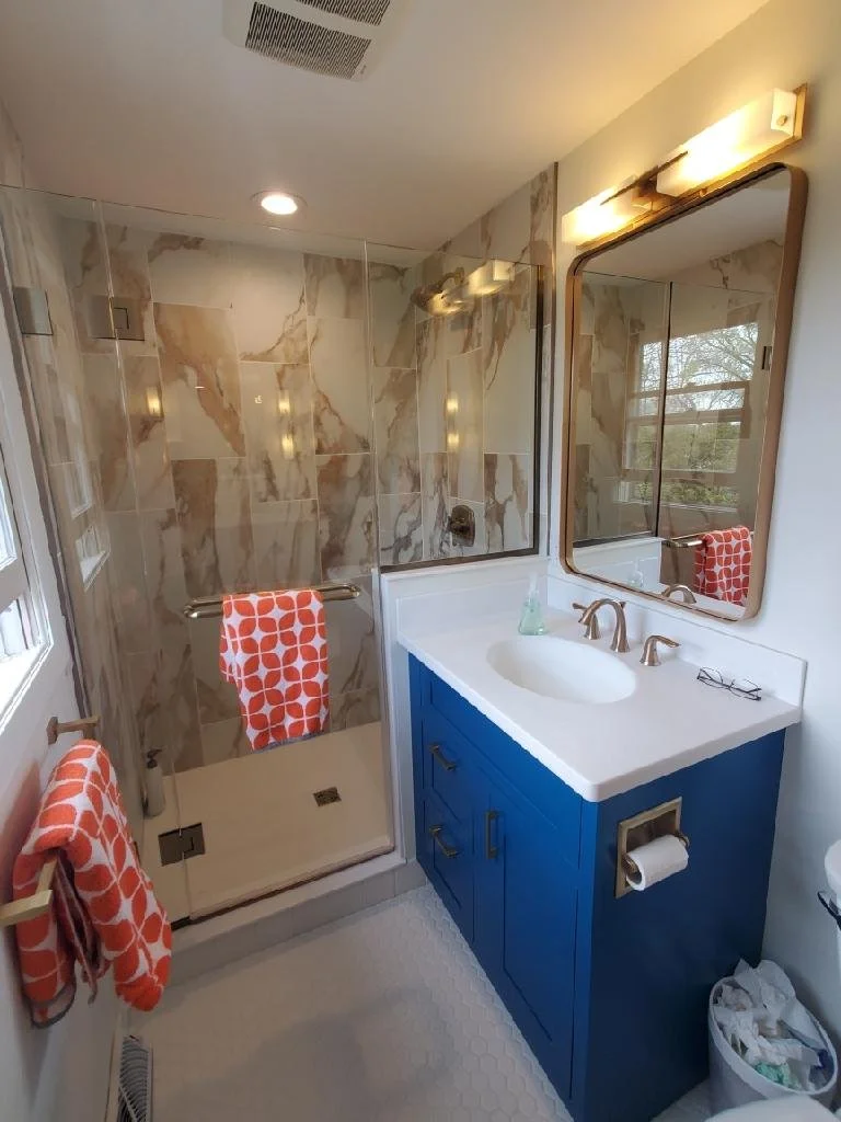 Bathroom with a glass-enclosed shower featuring marble tiles, a blue vanity with a white countertop and sink, a large mirror with warm lighting, and orange patterned towels.