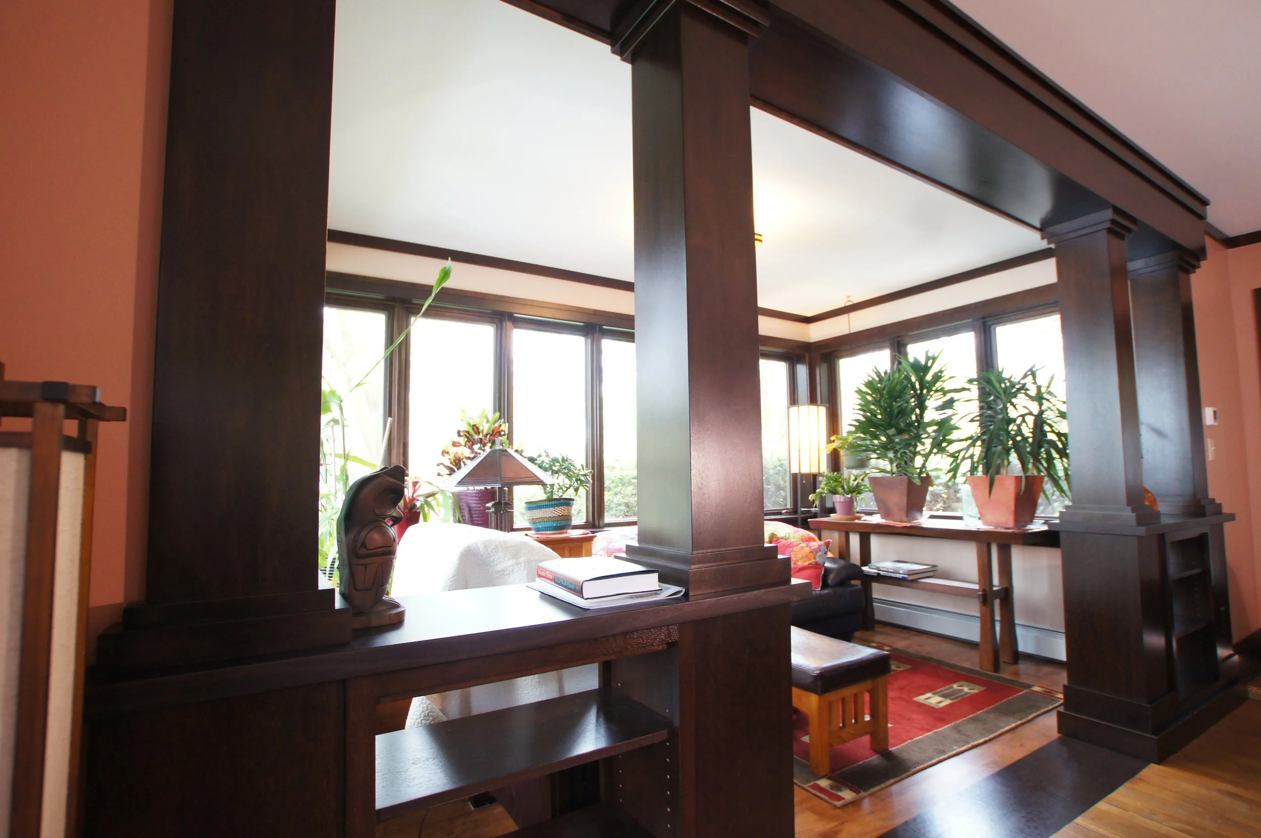 Living room with large windows, potted plants, a wooden partition, a lamp, books, and a rug on the hardwood floor.