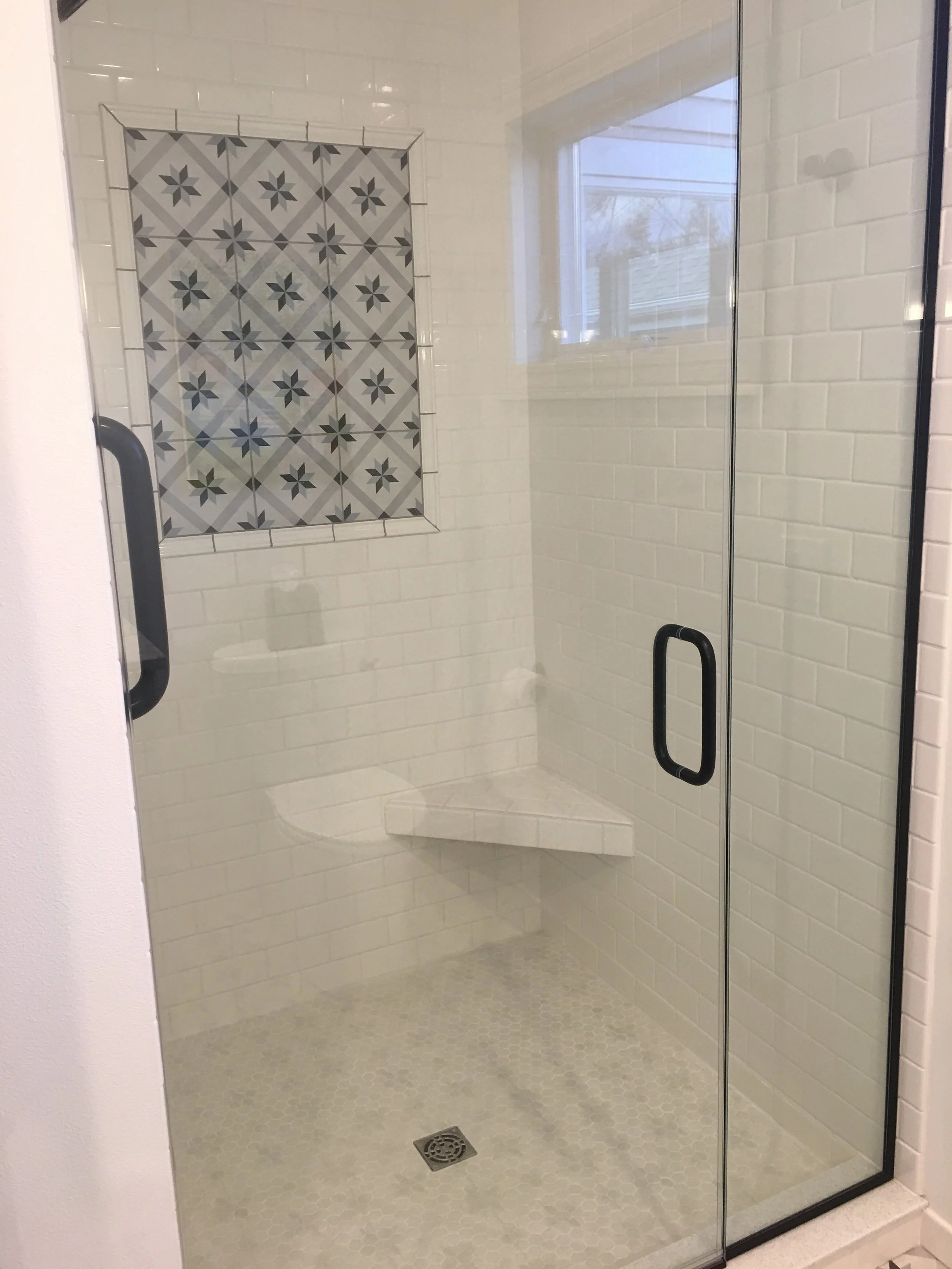 A modern walk-in shower with clear glass doors, white subway tile walls, a decorative tile accent with grey and white geometric and floral patterns, a built-in corner bench, and a small window near the ceiling.