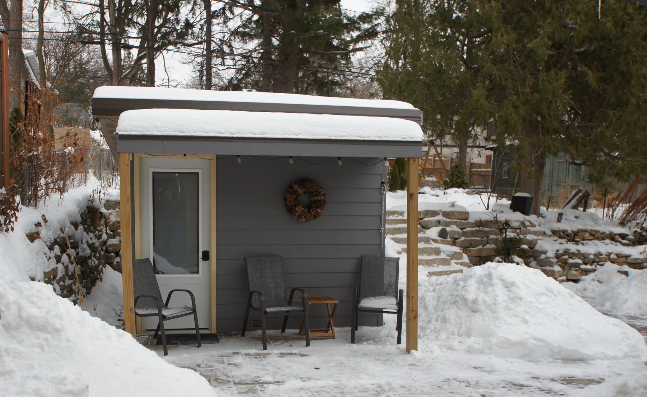 Small gray outdoor shed with snow on the roof, surrounded by snow-covered ground and a stone wall, with three gray chairs and a small wooden table in front, and a wintery backyard with trees and fencing in the background.