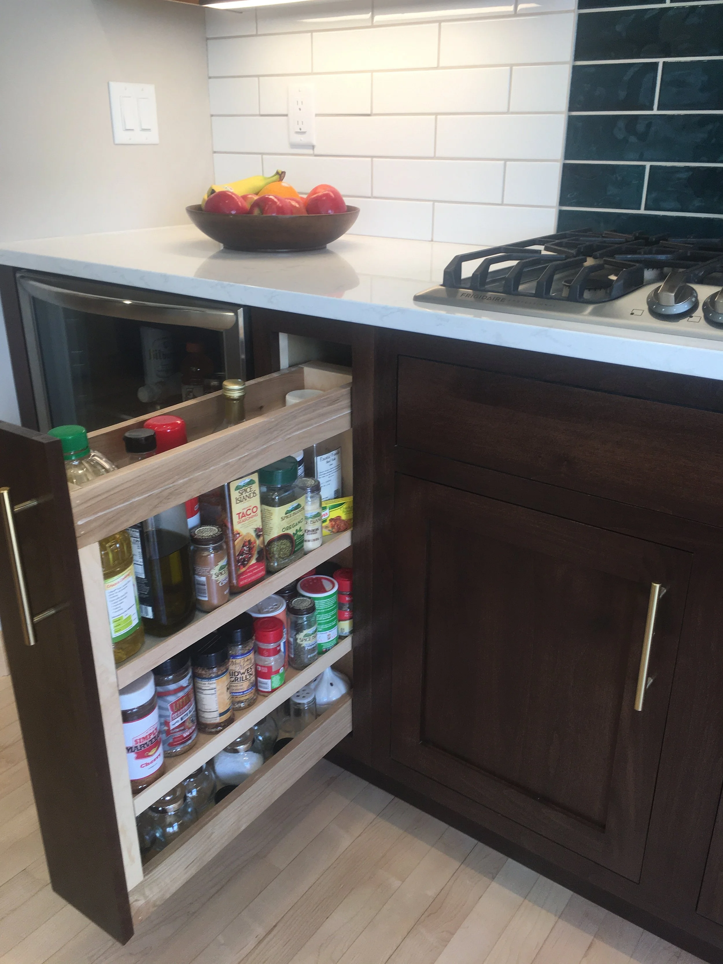 Open kitchen cabinet revealing spices and condiments, with a fruit bowl on the white countertop and a stove to the right.
