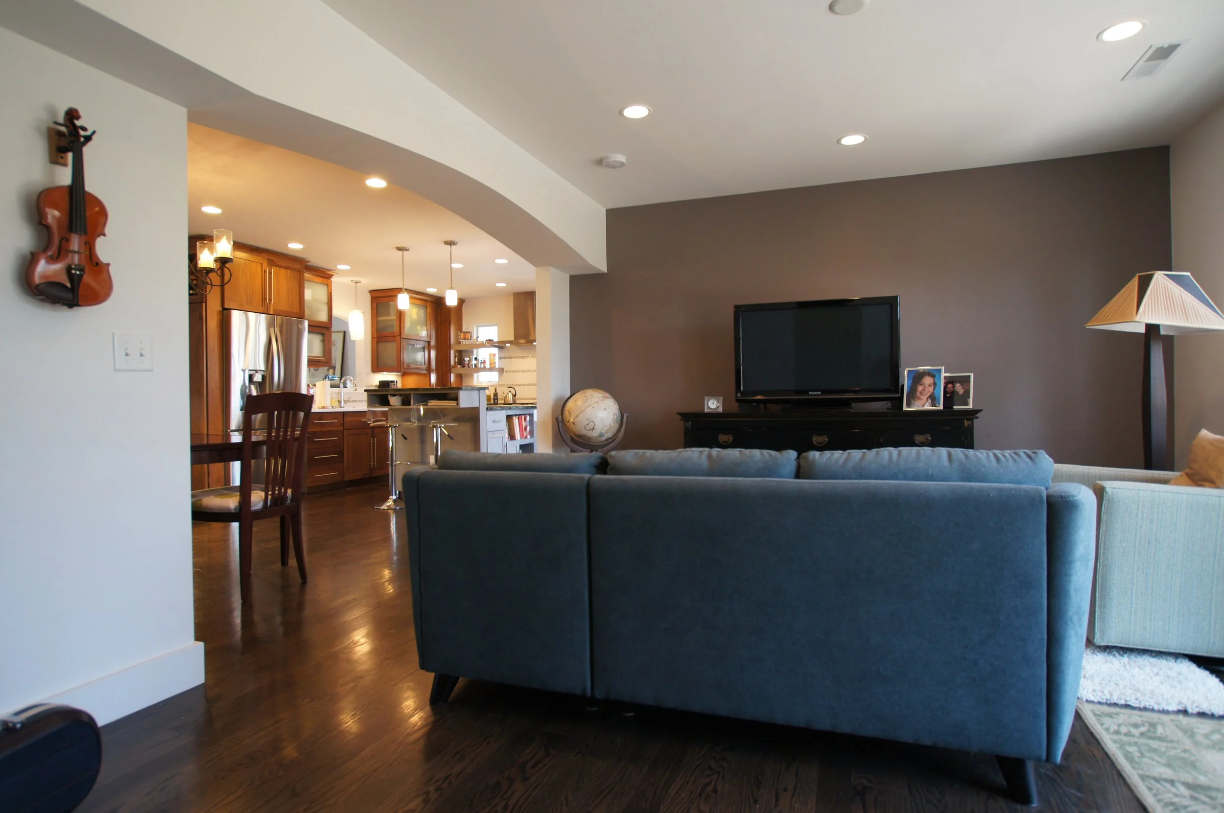 Living room with a blue couch, TV on a black stand, photograph frames, globe, lamp, and a dark brown wall, with an open kitchen in the background.