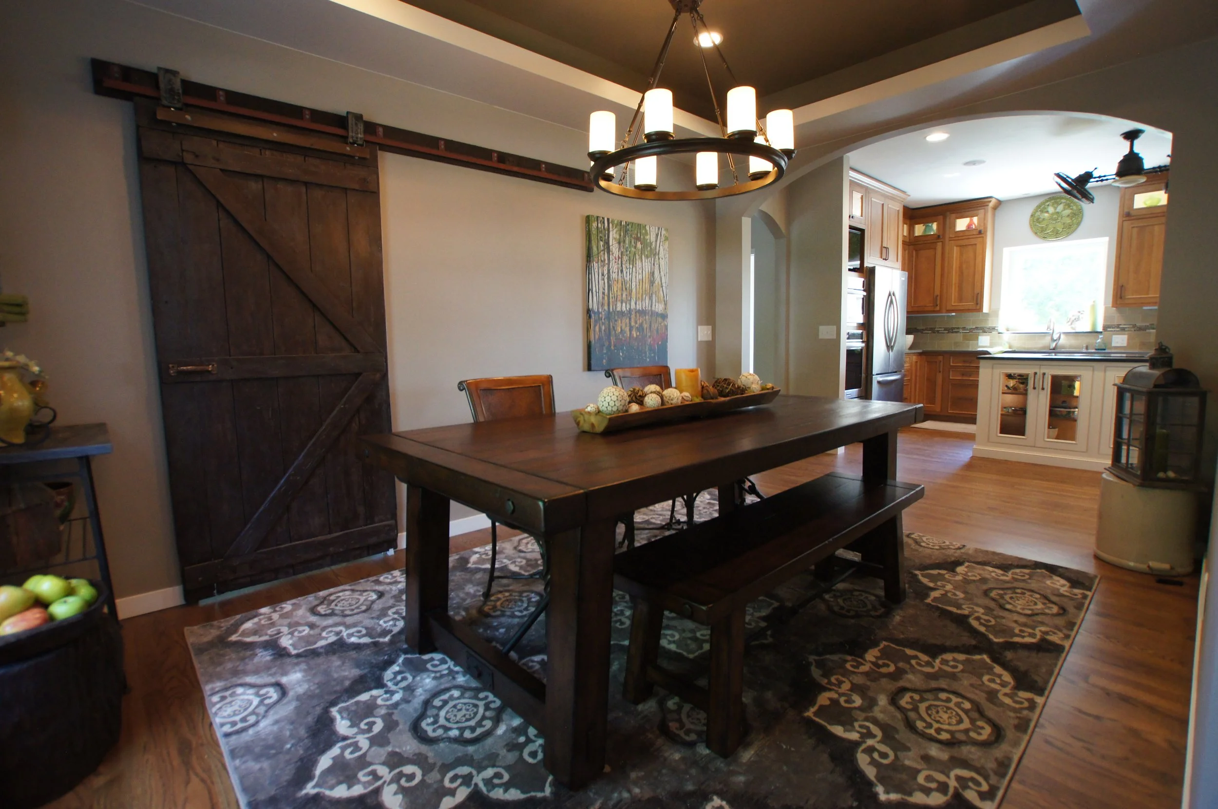 Dining room with wooden table, chairs, and bench, black chandelier, sliding barn door, and kitchen in the background with wooden cabinets and a window.