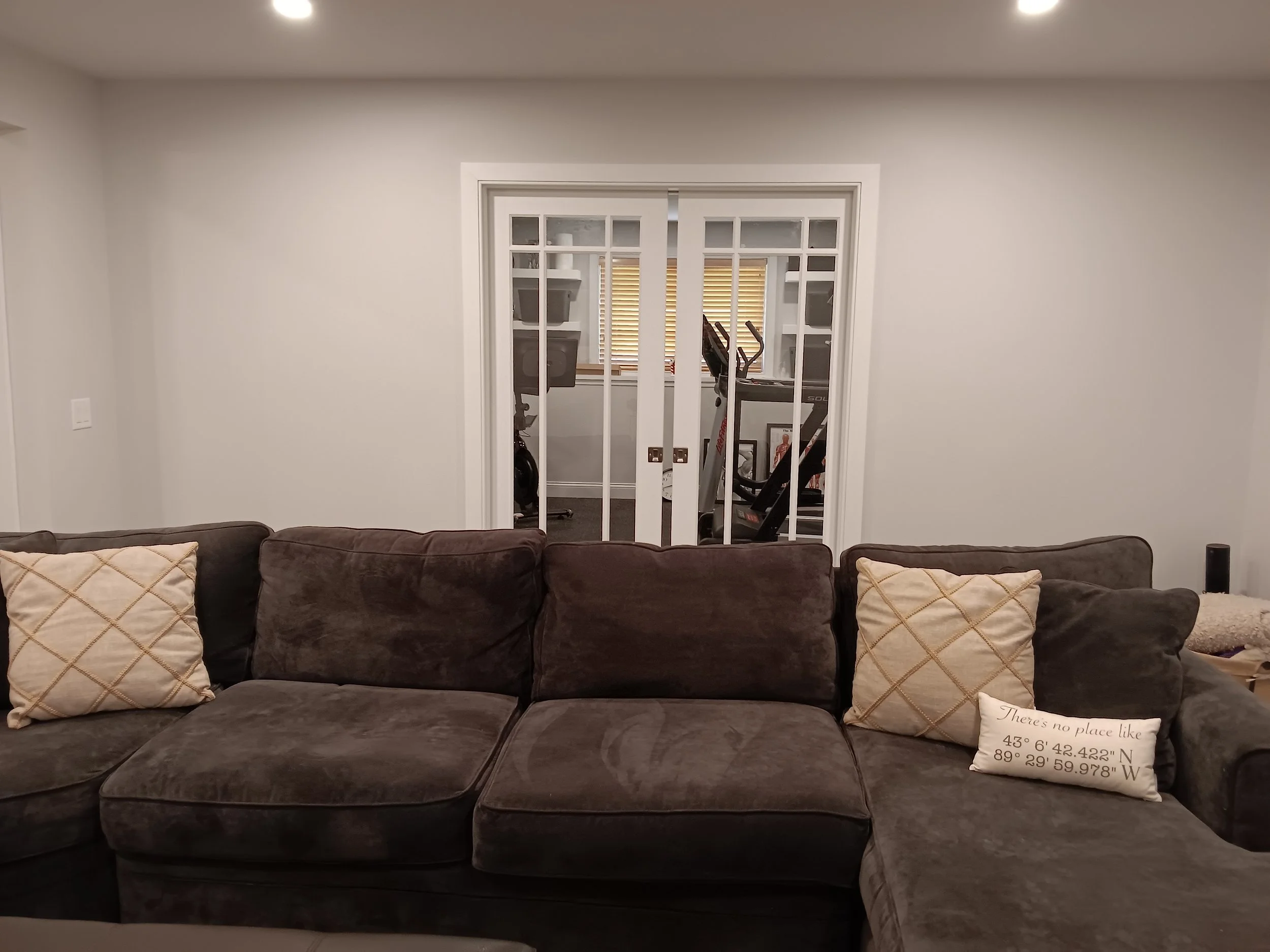 Living room with dark brown sectional sofa, beige pillows, and a sliding glass door leading to a room with exercise equipment.