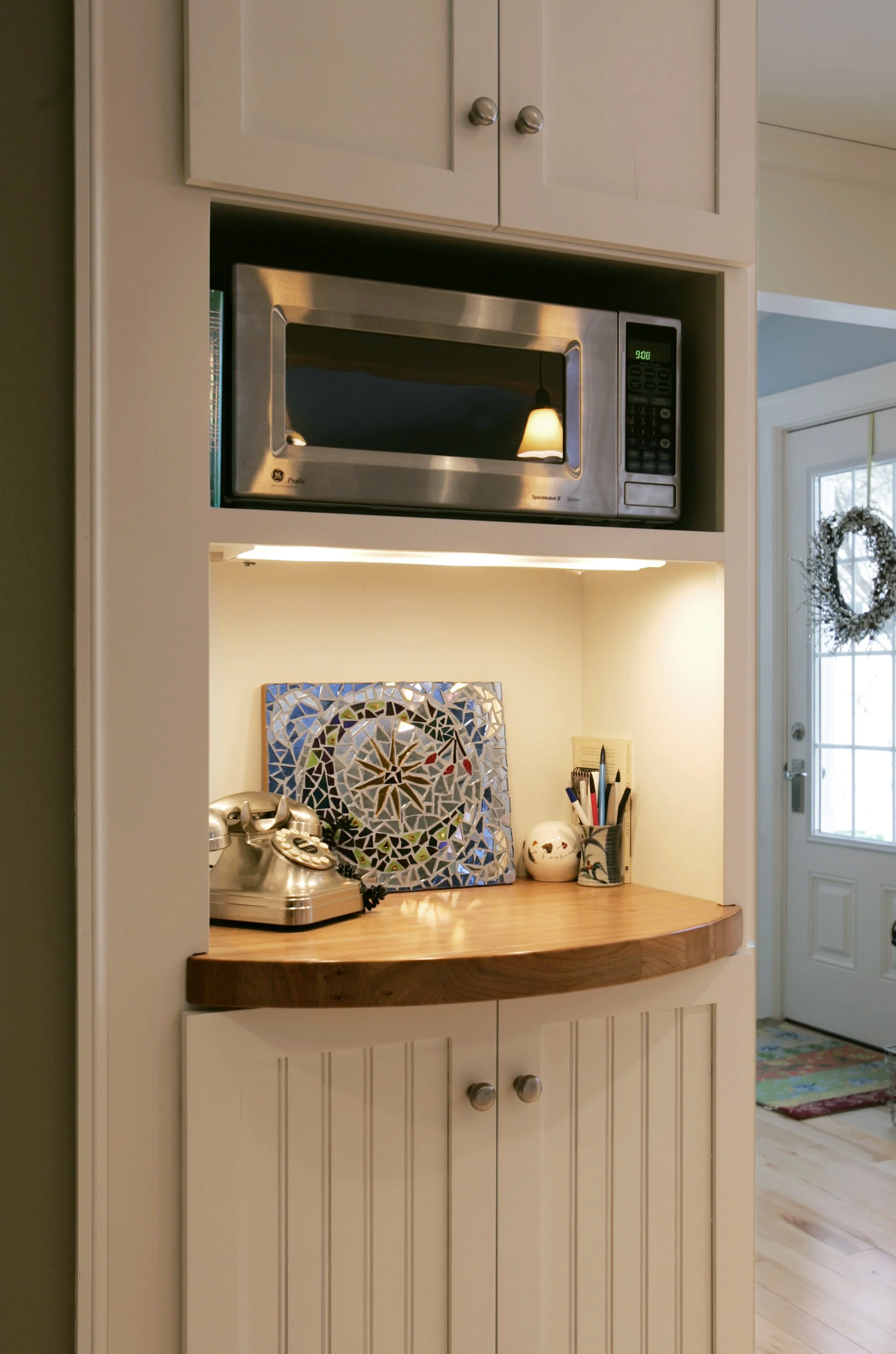 A built-in kitchen microwave above a corner countertop with a vintage rotary phone, a decorative mosaic piece, and a cup holding pens and pencils. A door with a holiday wreath is in the background.
