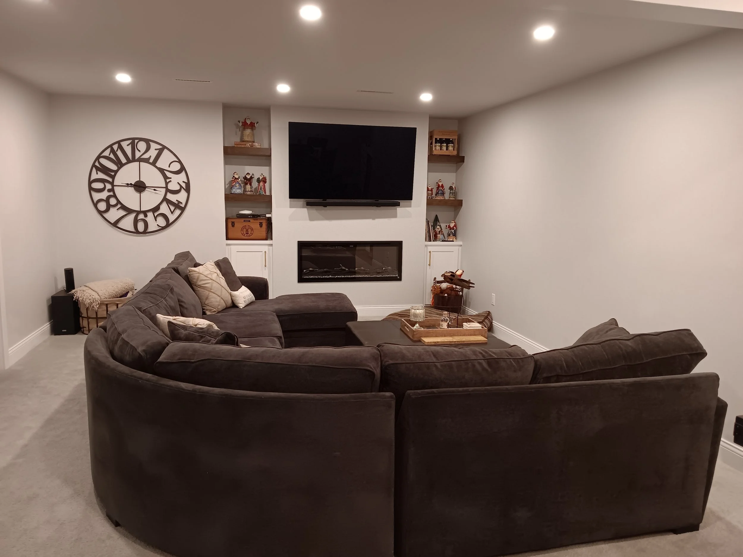 Living room with a large sectional sofa, mounted flat-screen TV, electric fireplace, decorative shelves with holiday figurines, and minimal wall decor.