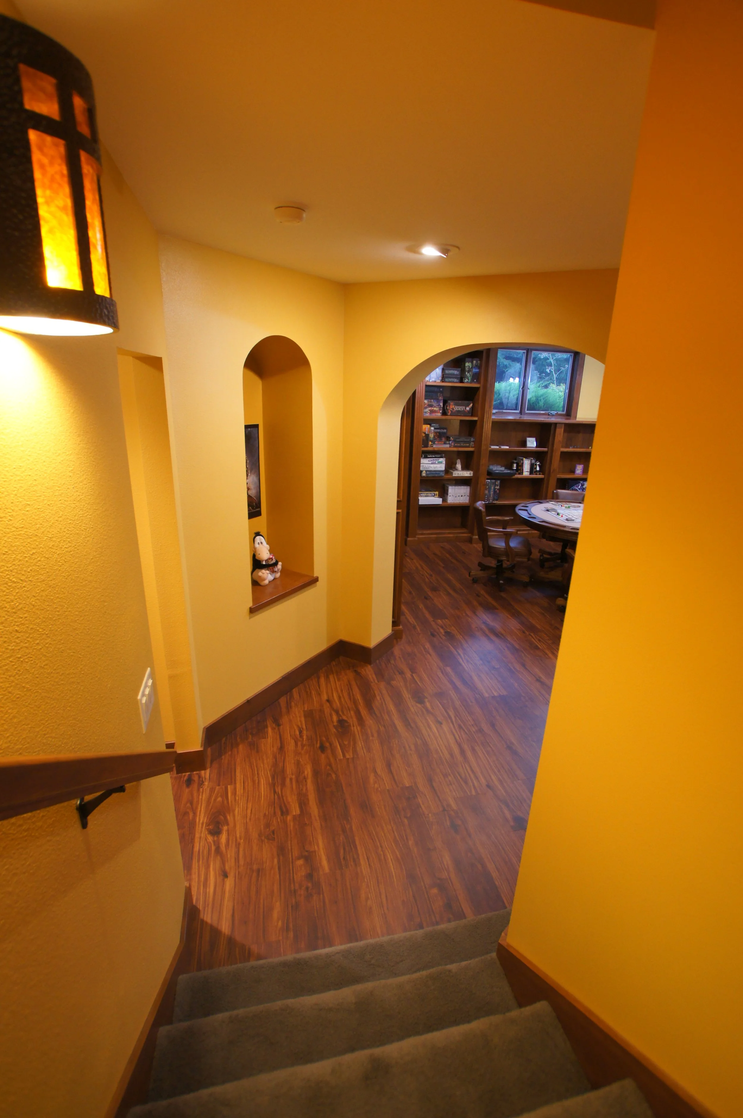 View of a hallway with yellow walls, wooden floors, and a staircase leading down. There is a small decorative shelf with a plush toy on a ledge, and in the background, a room with bookshelves, a round table, and chairs.