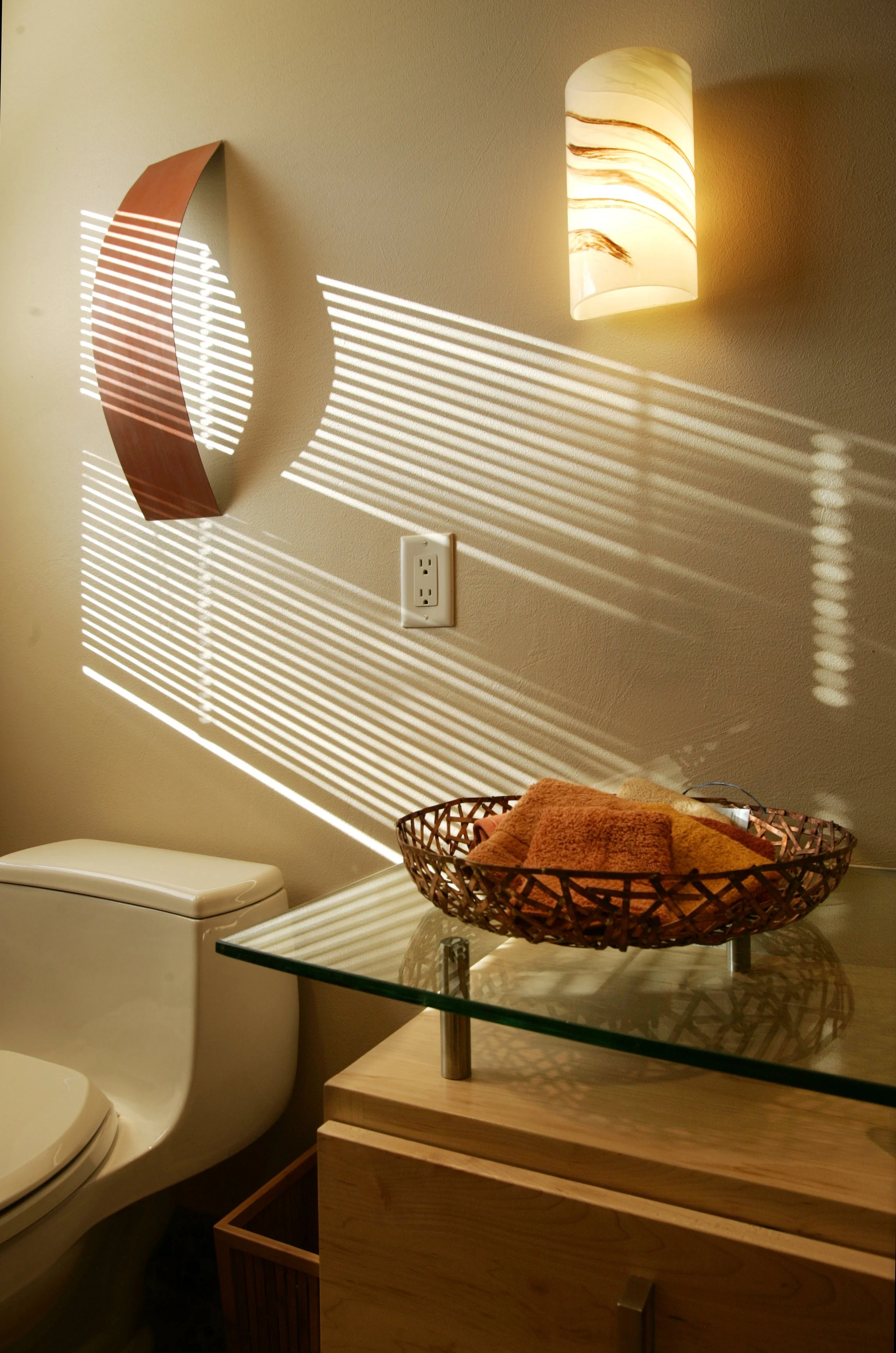 A bathroom corner with a beige wall, light fixture, a decorative wooden wall piece, a white electrical outlet, a glass-topped wooden cabinet with a basket of towels, and sunlight creating striped shadows on the wall.