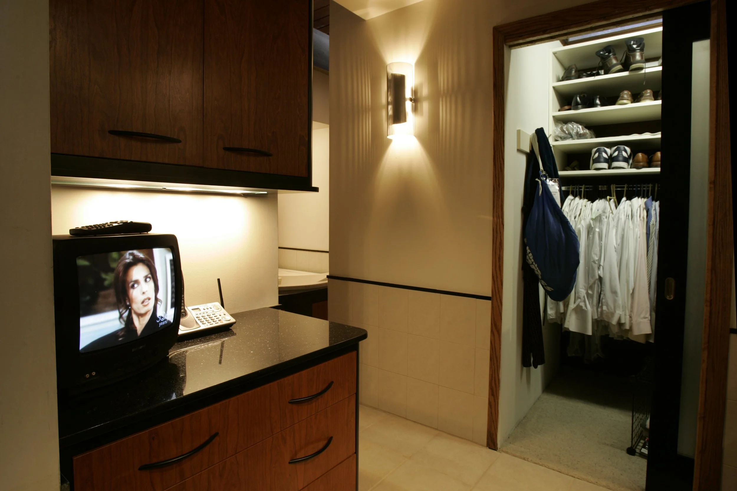 A hotel room with a small television on a black granite counter, showing a woman on screen, and a white landline phone beside it. There is an open closet with shoes on shelves and hanging clothes, partially visible on the right.