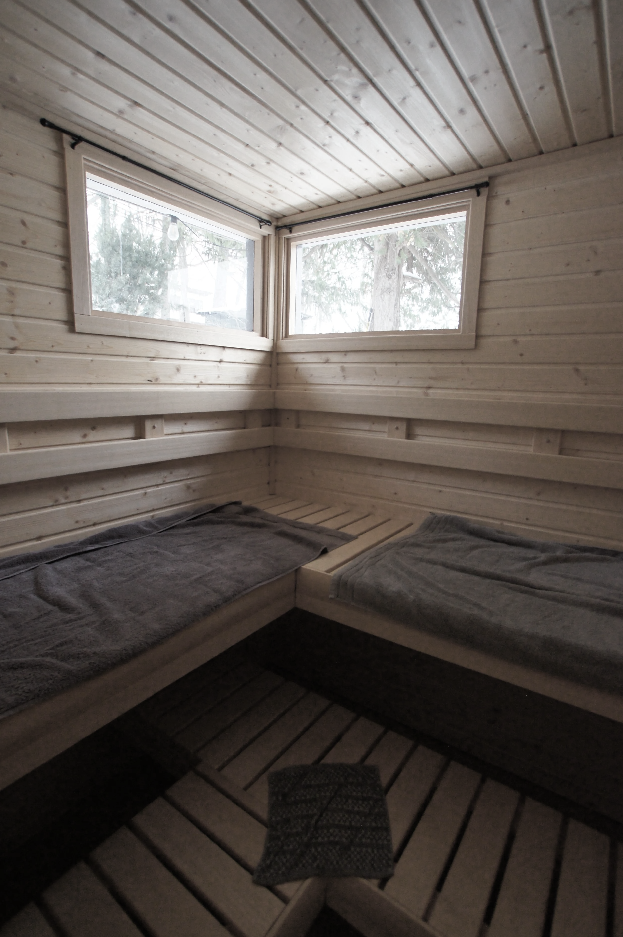 Interior of a small wooden room with two beds, each with folded towels, and windows showing trees outside.