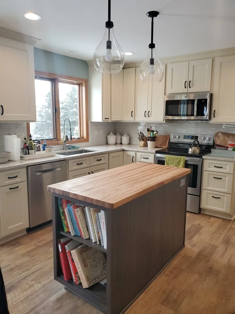 Kitchen with white cabinets, a countertop with kitchen items, a window, and a kitchen island with bookshelves.