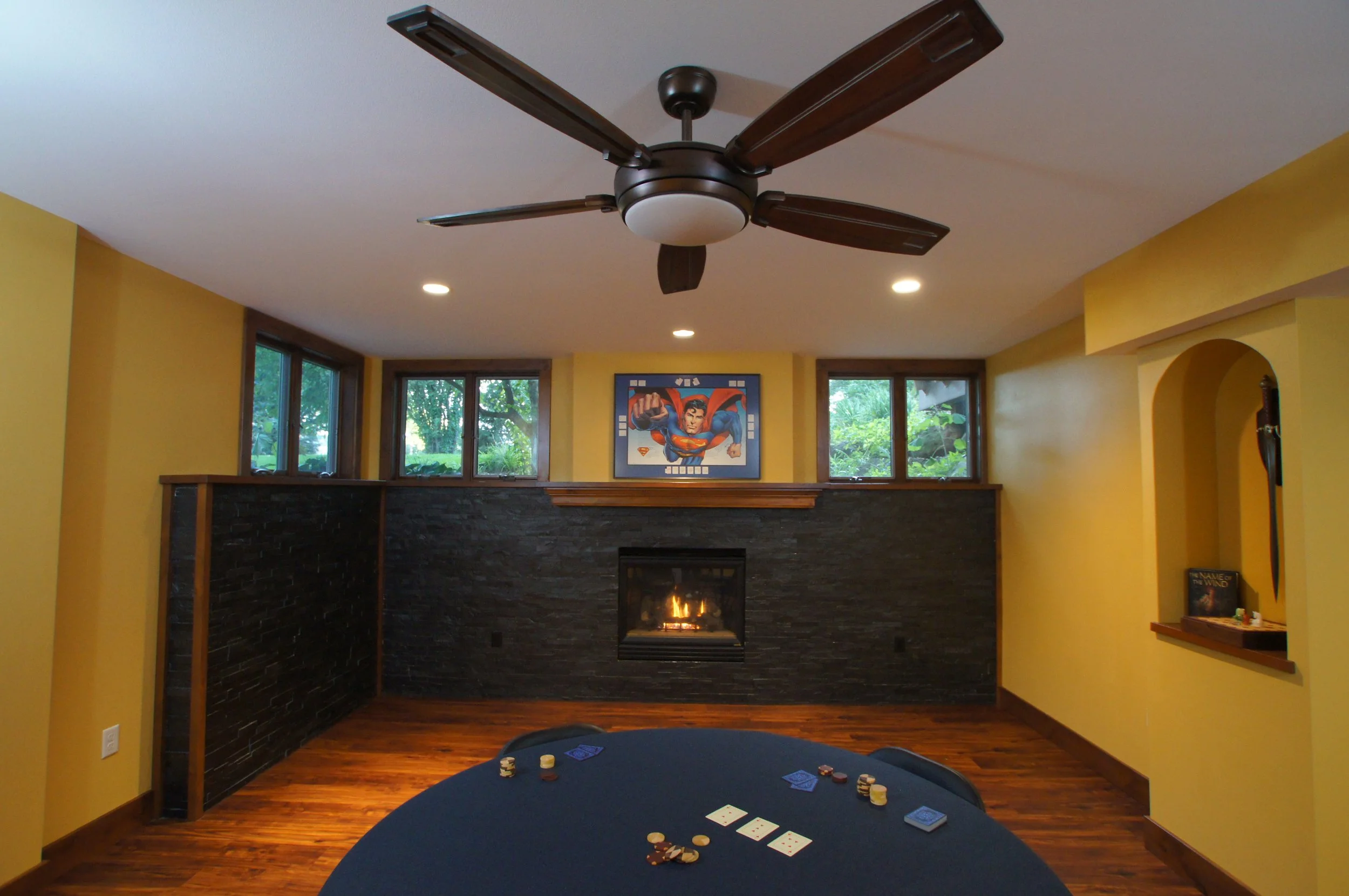 Living room with yellow walls, hardwood floor, dark stone fireplace, and three small windows above the fireplace. There is a ceiling fan with dark wood blades and a light fixture. A framed Superman artwork is hung above the fireplace. In the foregrou
