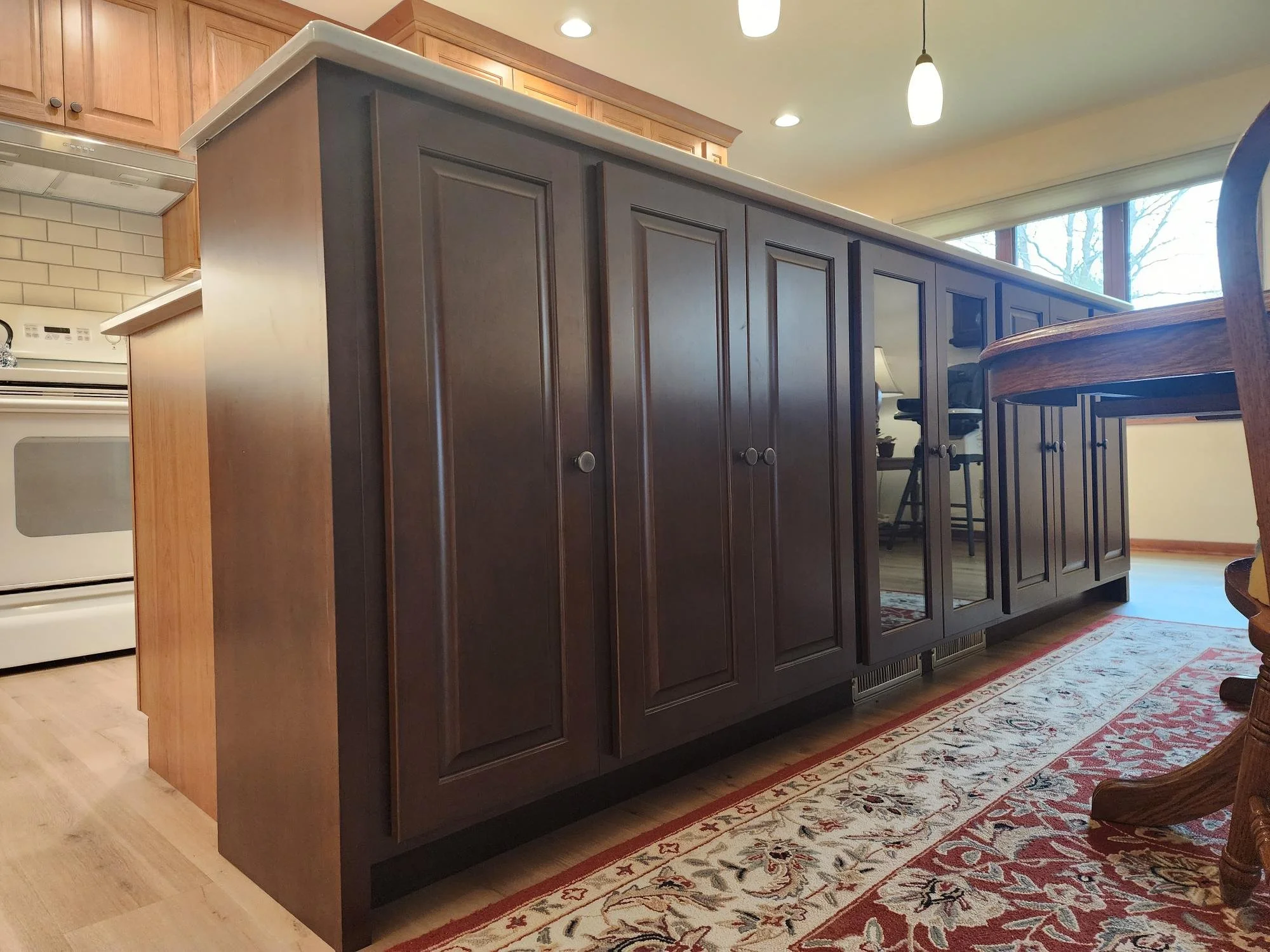 A kitchen island with dark wood cabinets and a white countertop, with a window and wooden chairs nearby.