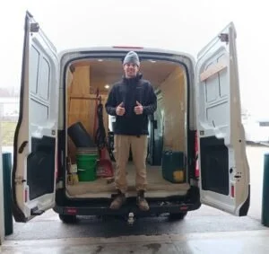 Man standing in the open back of a cargo van, which is parked indoors, with various tools and supplies inside.