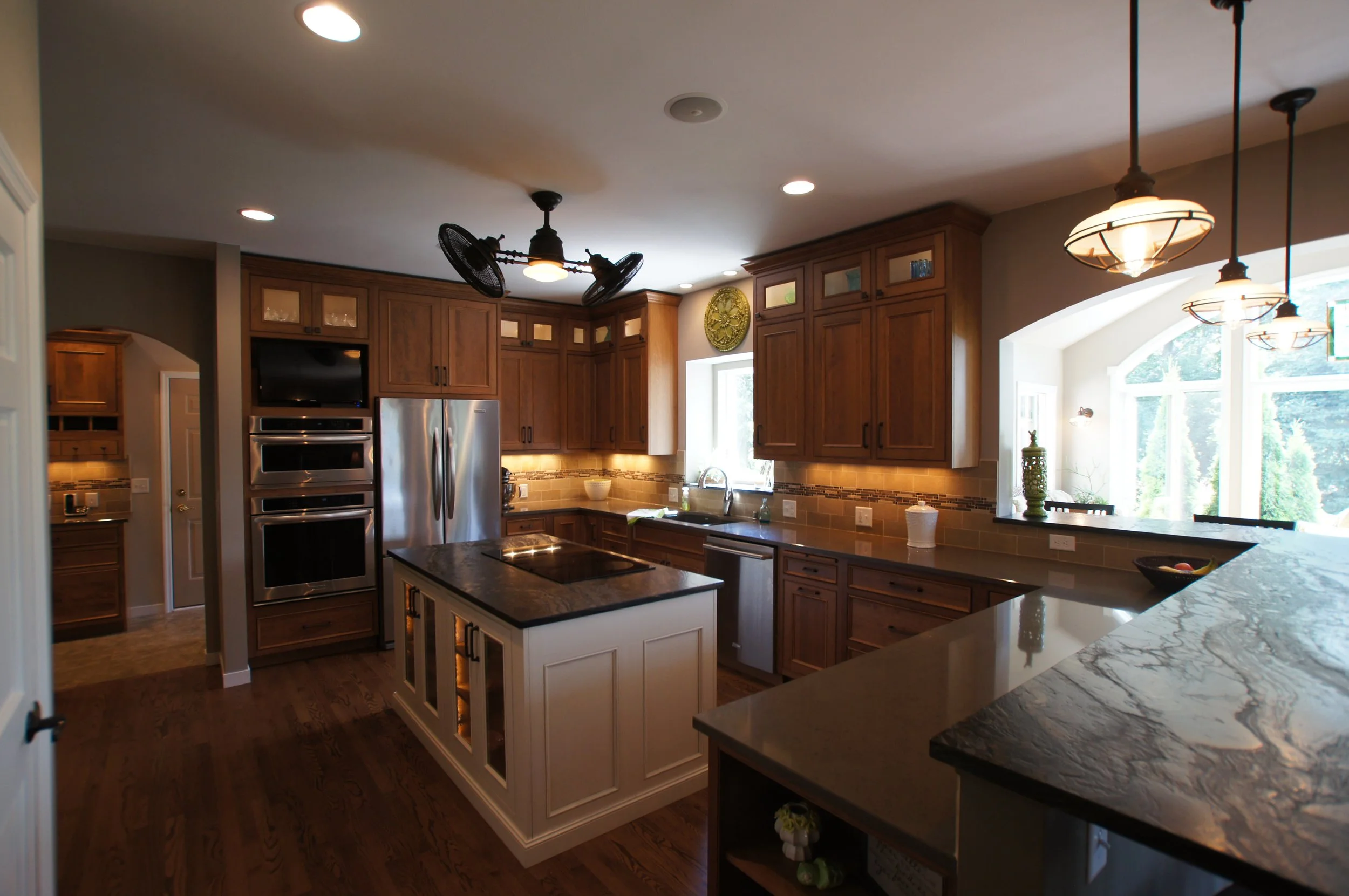 A spacious kitchen with wooden cabinets, a center island with a dark granite countertop, stainless steel appliances, pendant and ceiling lights, and large windows letting in natural light.