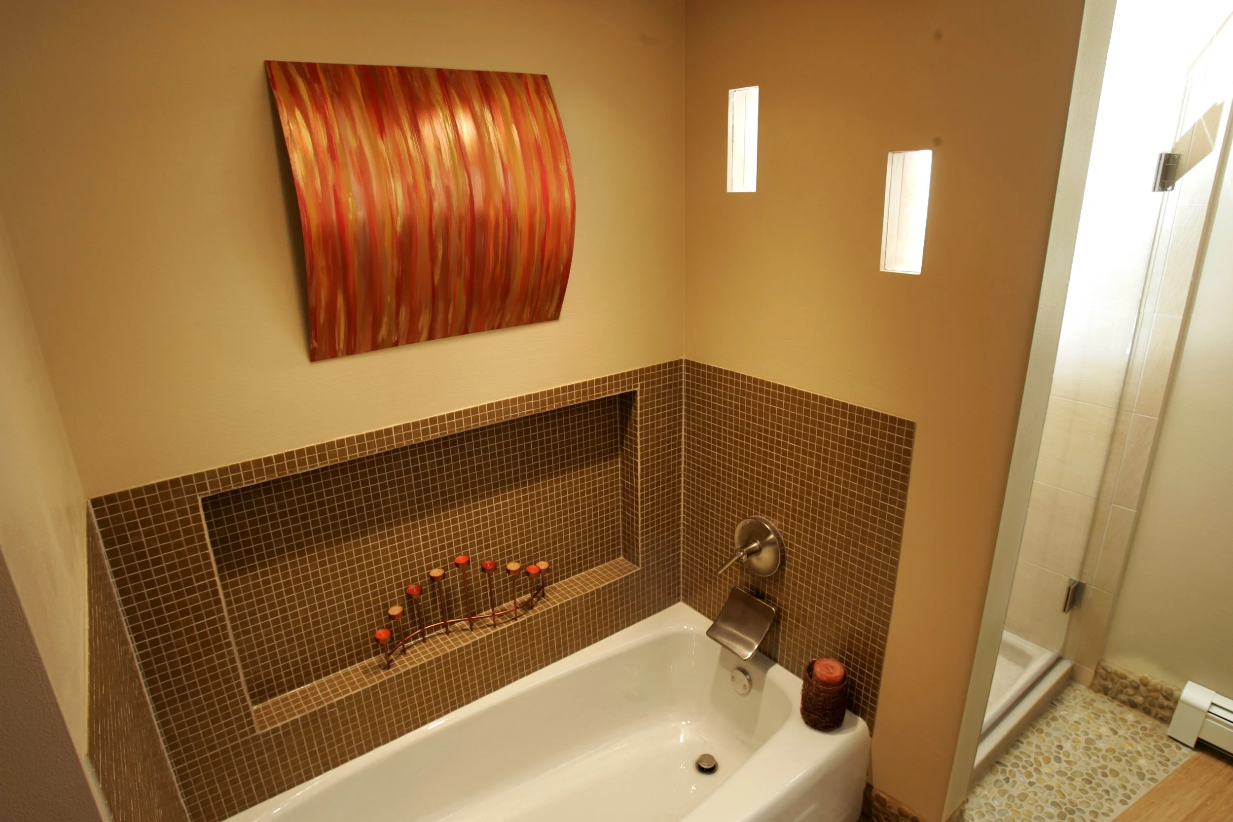 Bathroom with a white bathtub, brown mosaic tile wall, decorative candle, and a shower stall with pebble floor, three small windows, and artwork on the wall.