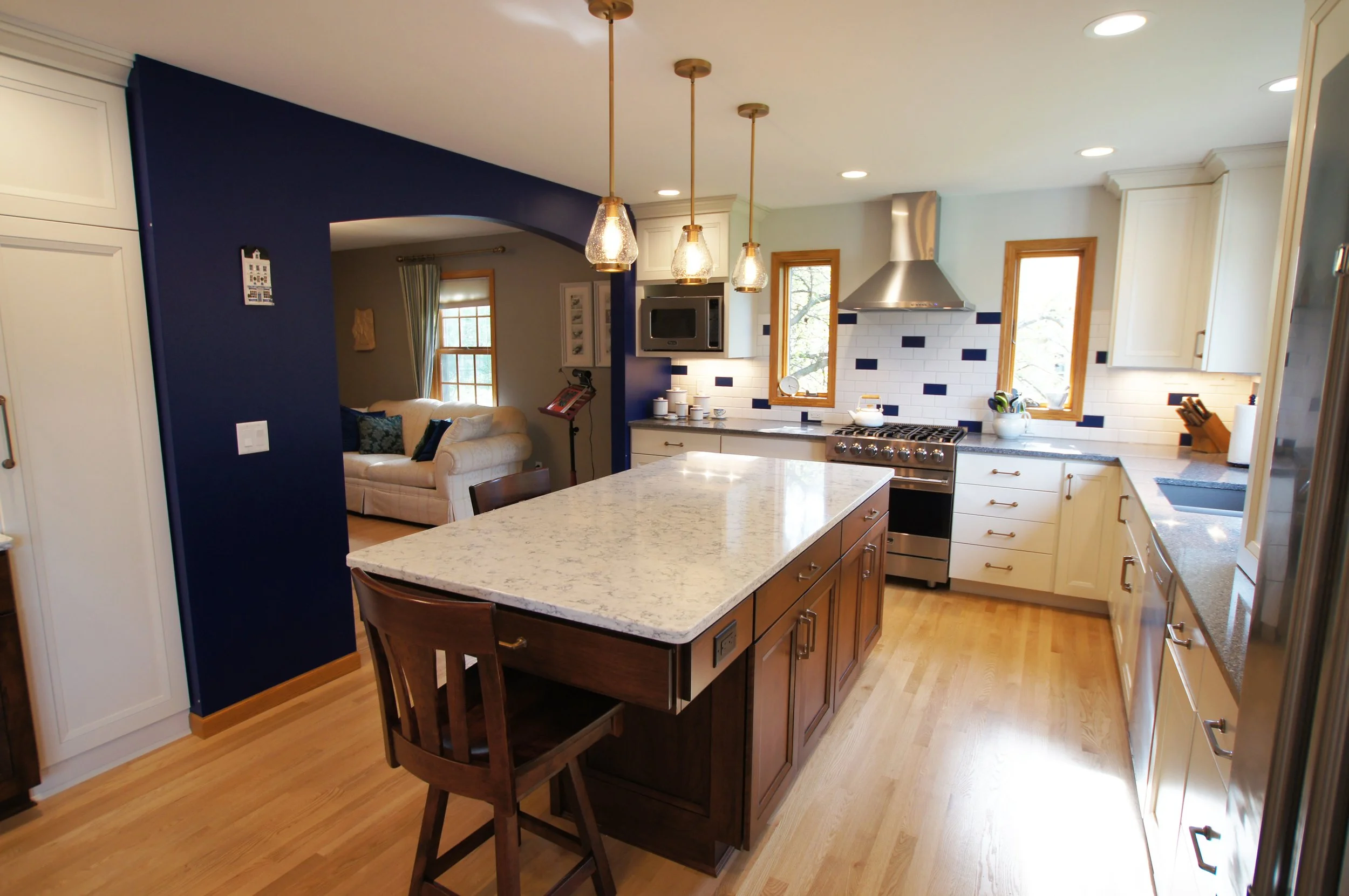 Modern kitchen with white cabinets, stainless steel appliances, and a large granite island. Blue accent wall, three pendant lights, and wooden floors.