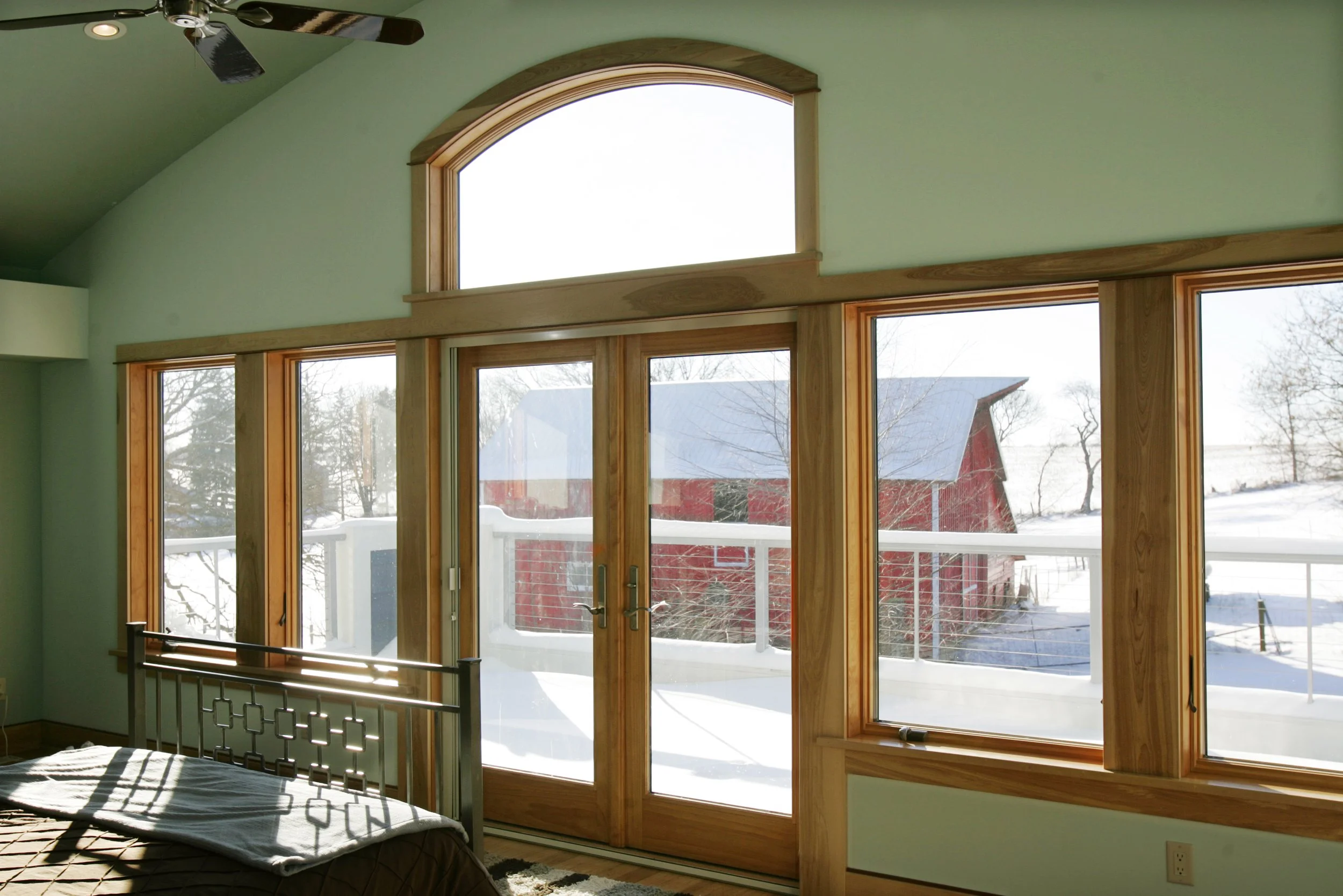 Interior of a room with large wooden-framed windows and a sliding glass door overlooking a snowy outdoor landscape with a red building and leafless trees, sunlight casting shadows inside.