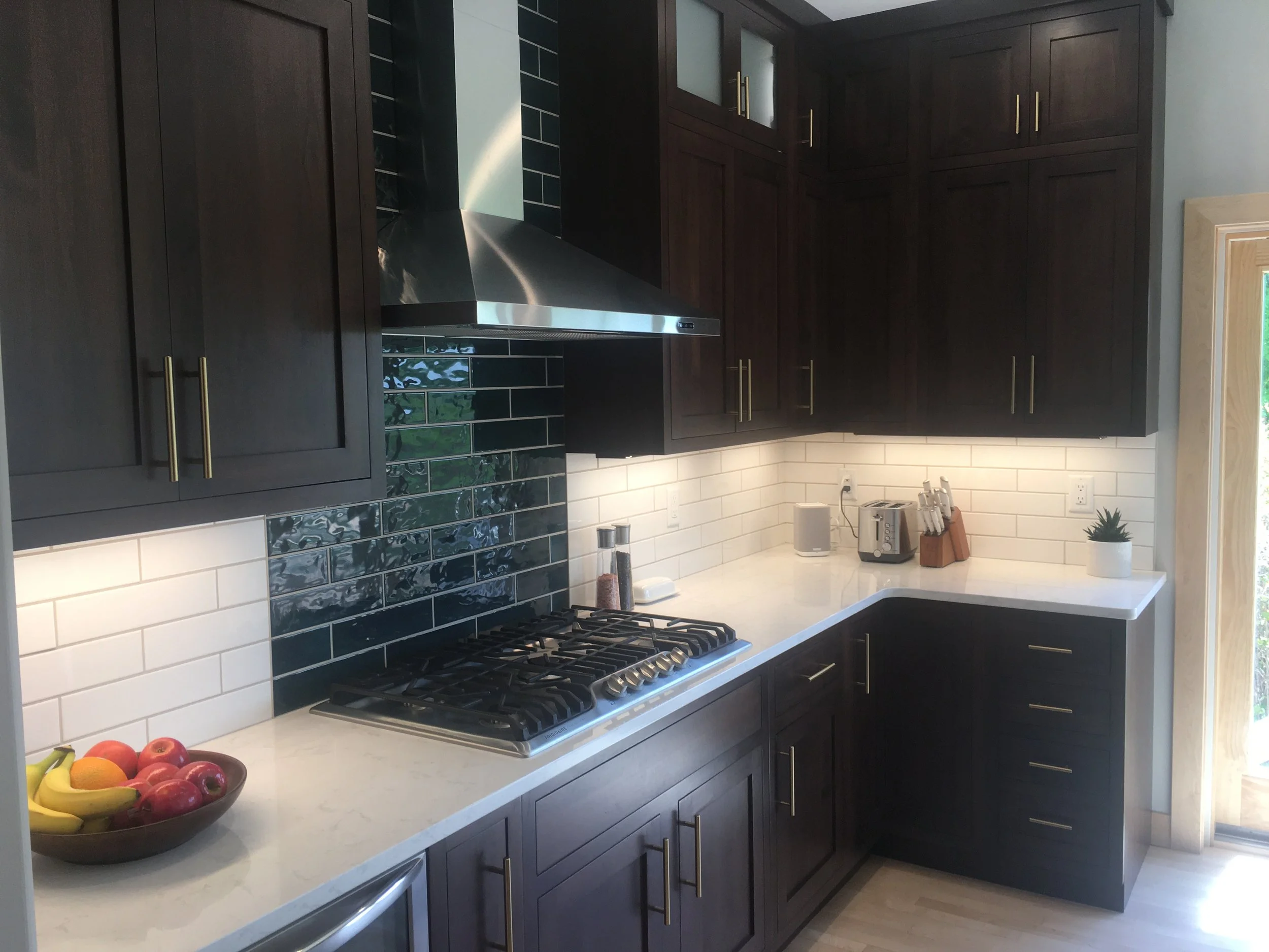 Modern kitchen with dark wood cabinets, white countertops, black and white tiled backsplash, stainless steel range hood, and small appliances on the counter.