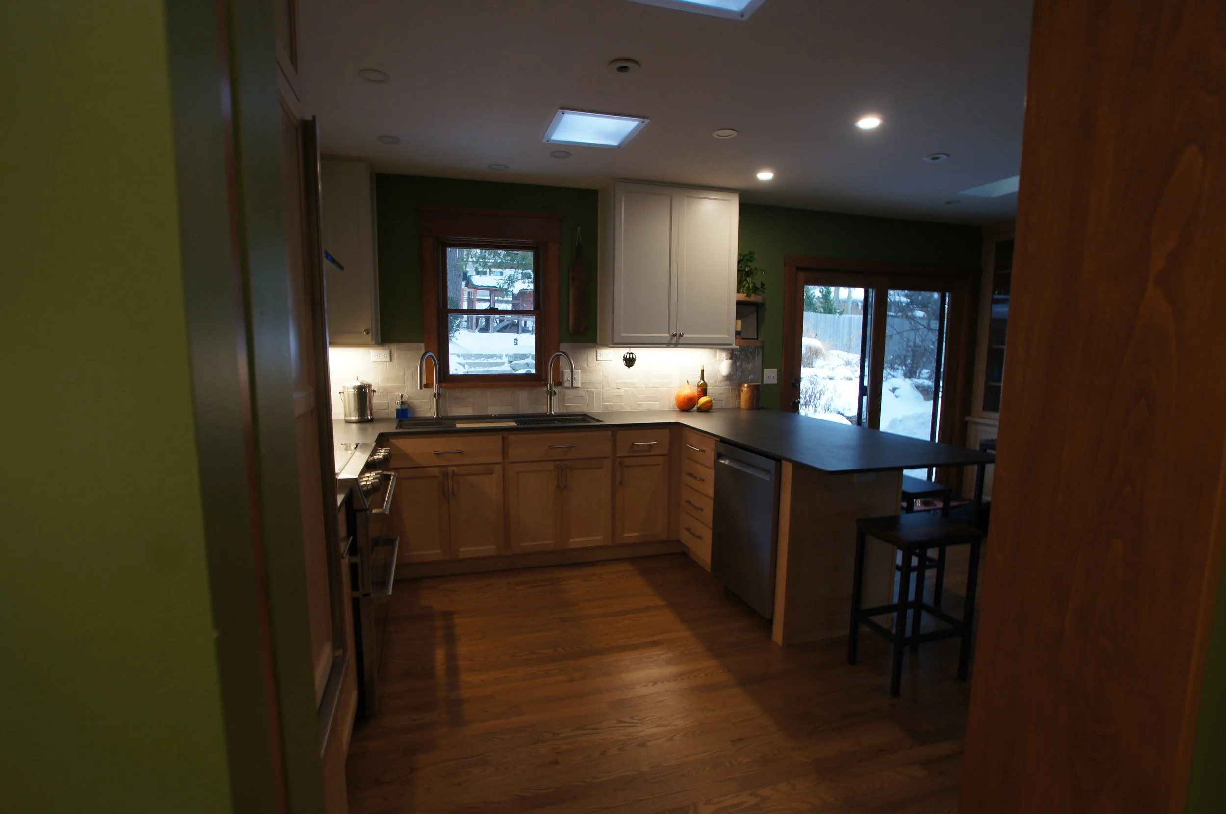 A cozy kitchen with wooden cabinets, a black countertop, and a view of a snowy backyard through windows. The kitchen is lit with recessed ceiling lights and features a double sink under a window, with a small island and bar stools adjacent to a slidi