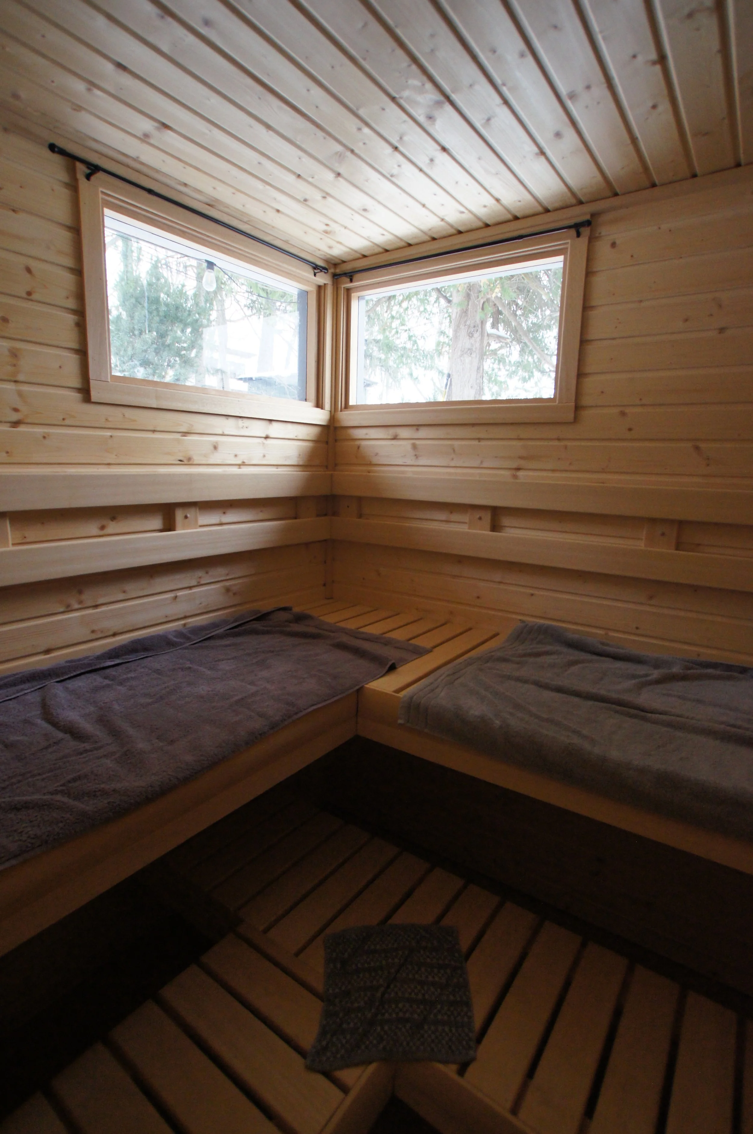 A wooden cabin bedroom with two beds, each covered with dark towels, and two large windows showing trees outside.