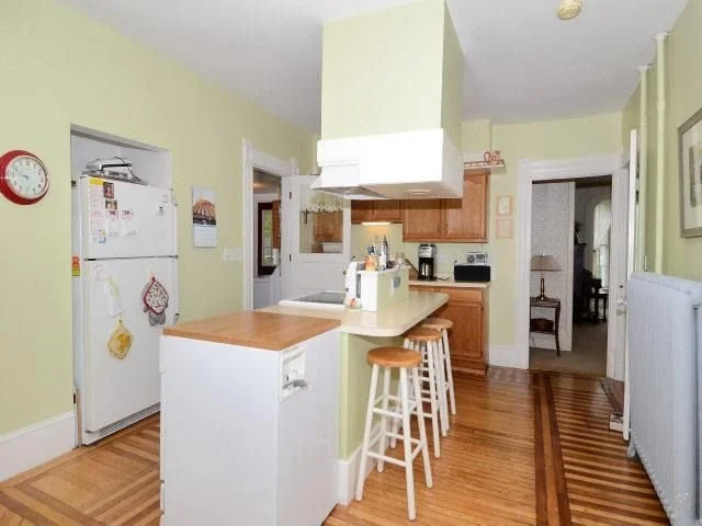 Kitchen with light green walls, white refrigerator, wooden cabinets, small island with chairs, and hardwood floors.
