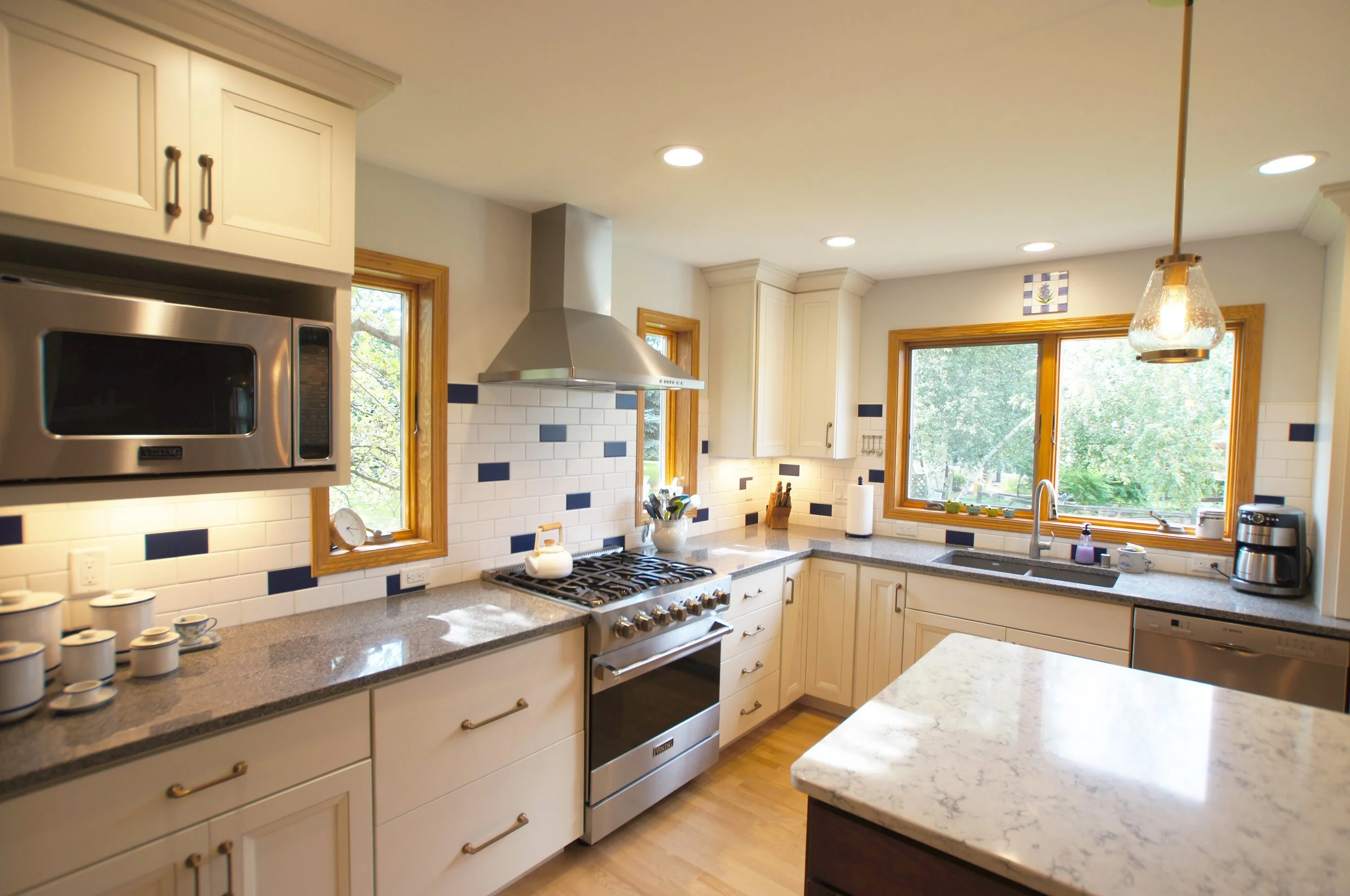 Bright kitchen with white cabinets, granite countertops, and large windows showing green trees outside. Includes stainless steel stove, microwave, and coffee maker. Tiled backsplash with white and navy blue tiles, and pendant light over the island.