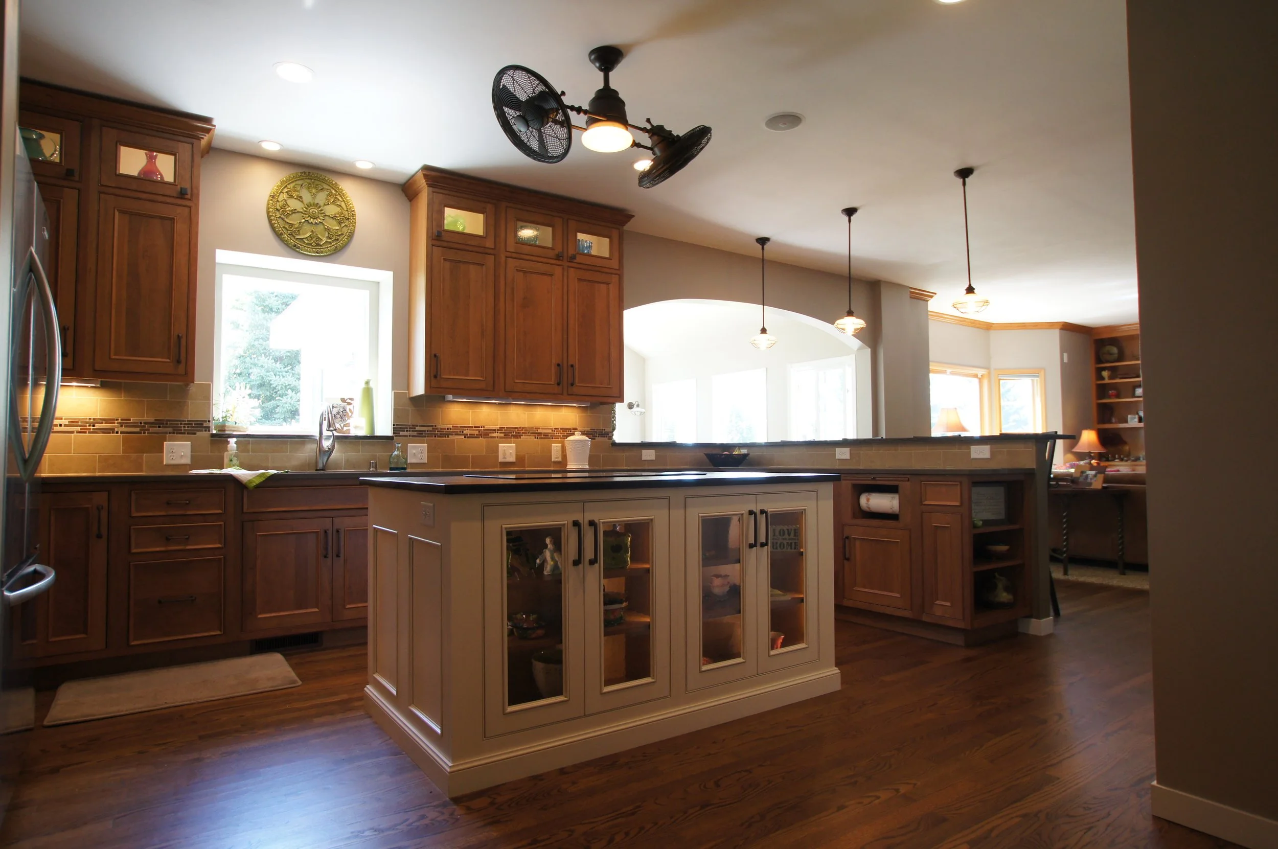 Kitchen with wooden cabinets, a central island with glass-front cabinets, hanging pendant lights, and a ceiling fan, with a window above the sink.