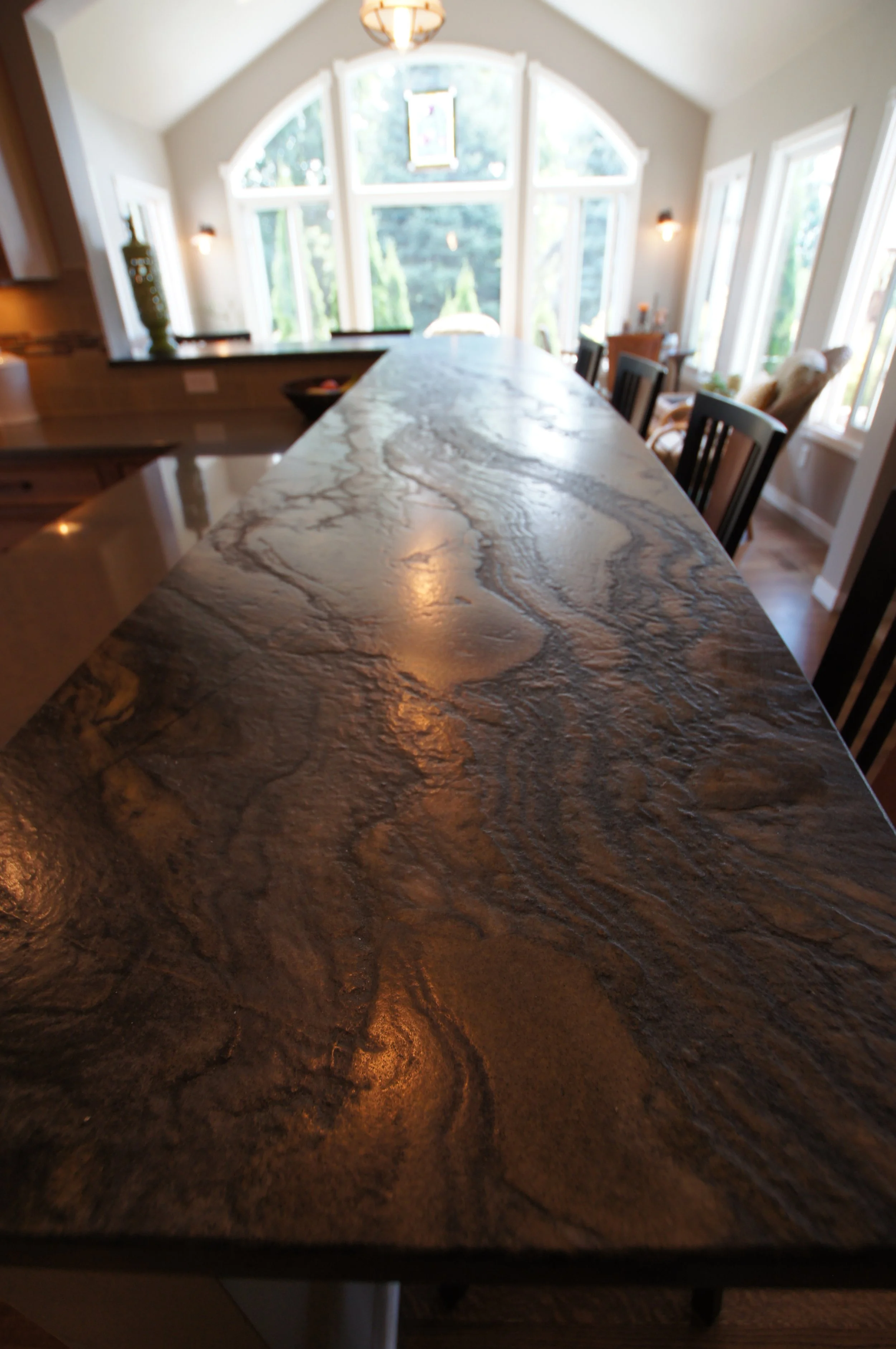 Close-up of a polished granite kitchen island or countertop with natural swirling patterns, set in a bright kitchen with large windows and dining chairs.