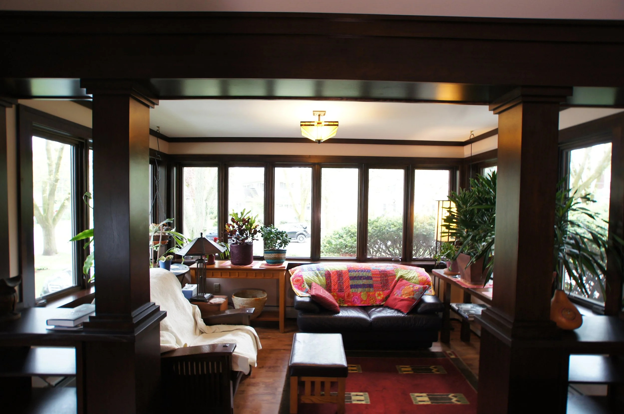 Living room with large windows, dark wood trim, a black sofa with colorful throw pillows, wooden furniture, and indoor plants.