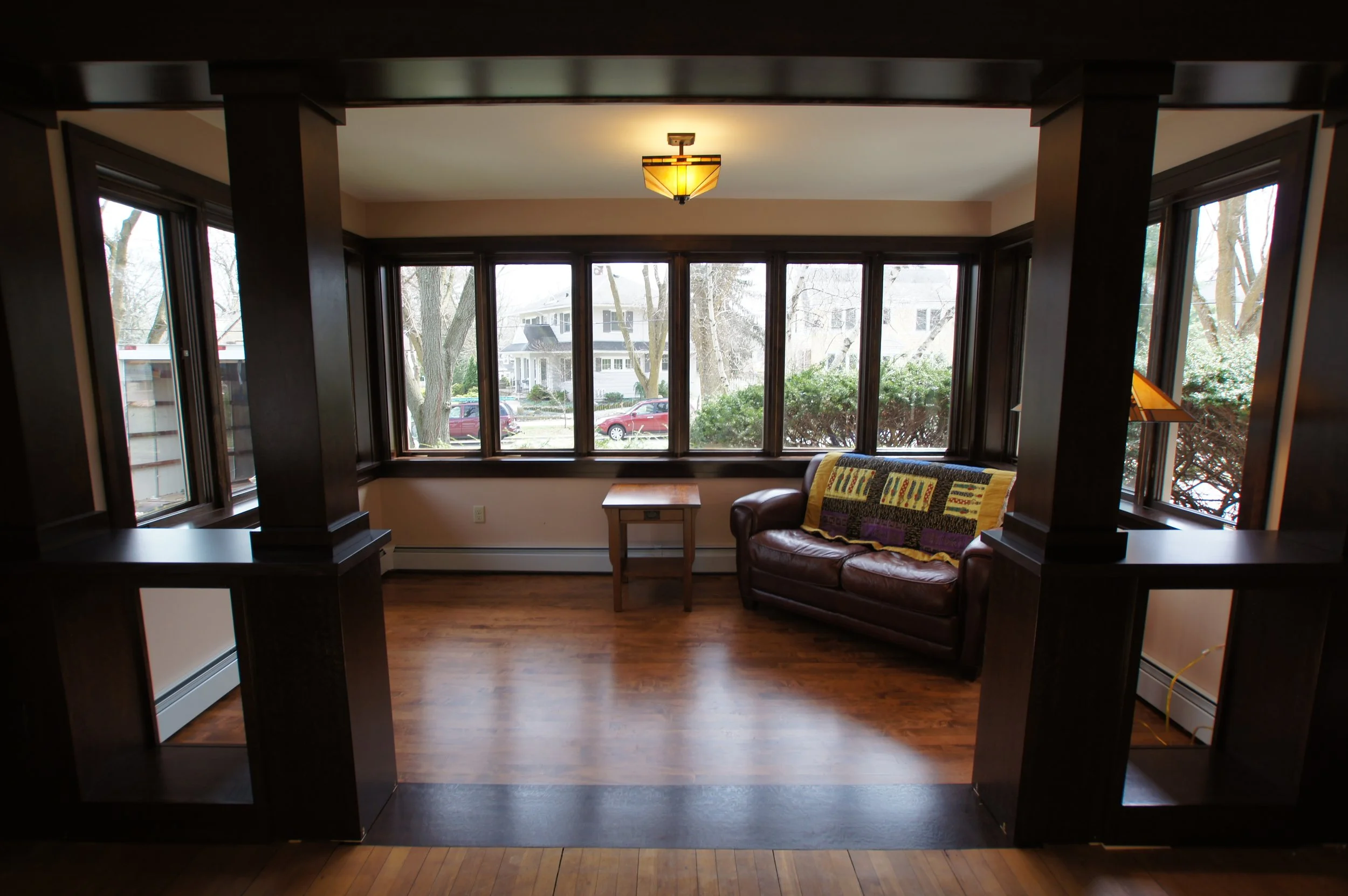 View of a living room with large bay windows, a brown leather couch with a colorful quilt, a small wooden table, and hardwood floors, with trees and houses visible outside.