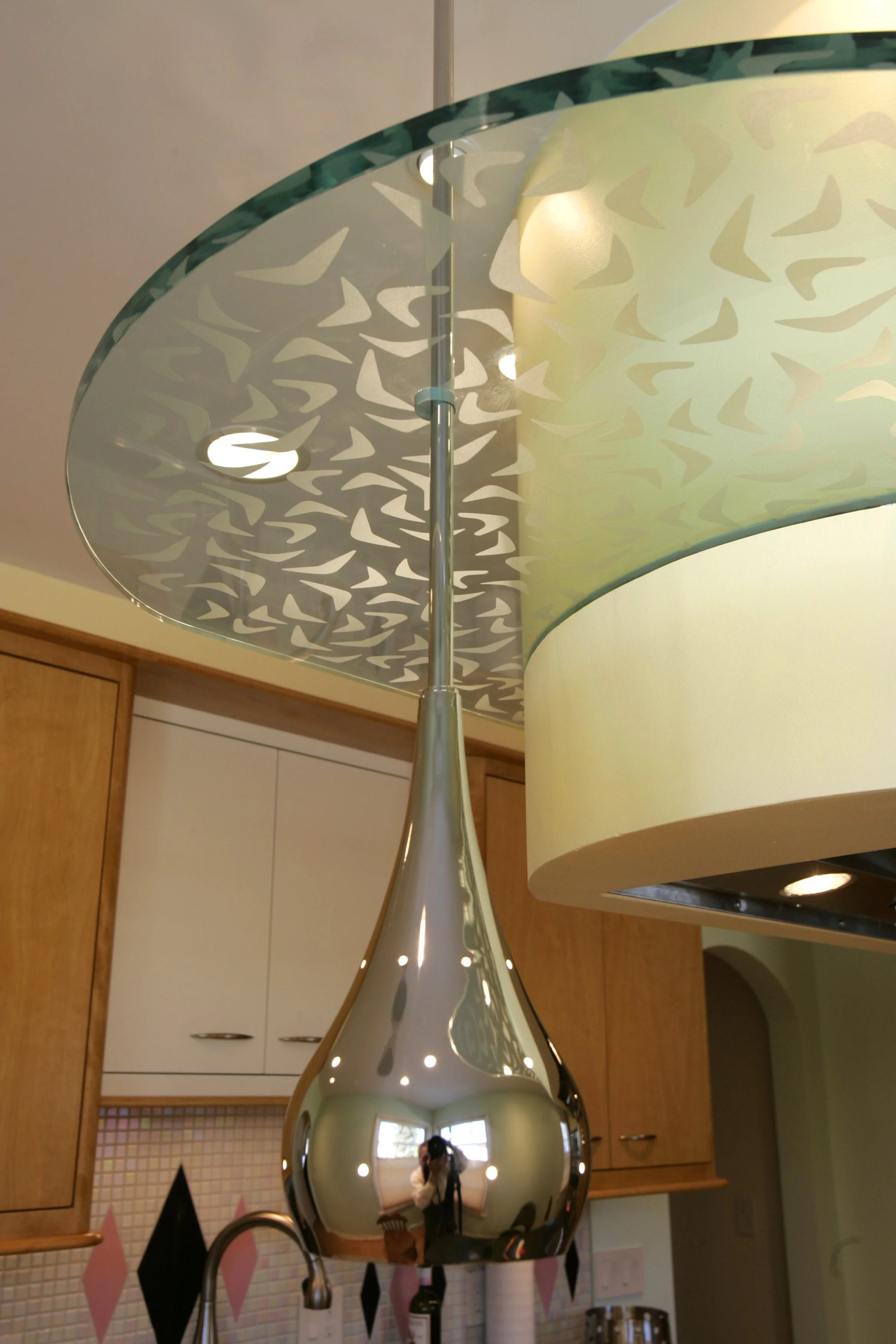 Close-up view of a modern kitchen light fixture with a reflective metallic teardrop-shaped pendant hanging from a decorative glass cover with abstract cut-out design, mounted above a kitchen sink.