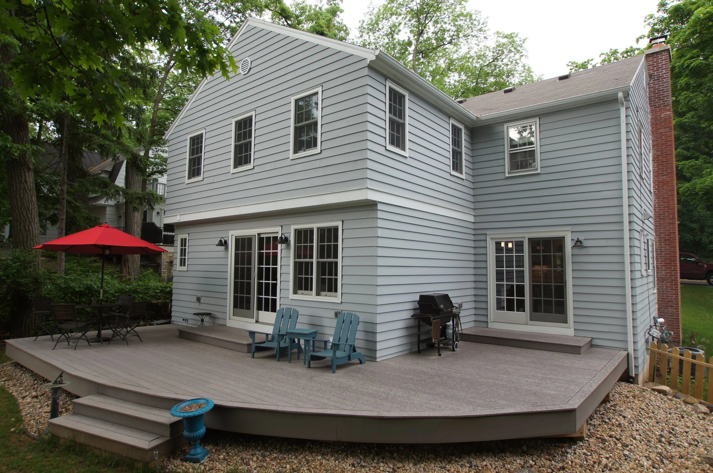 Backyard deck attached to a gray house with multiple windows and glass doors, outdoor furniture, a red patio umbrella, a grill, and surrounding trees.