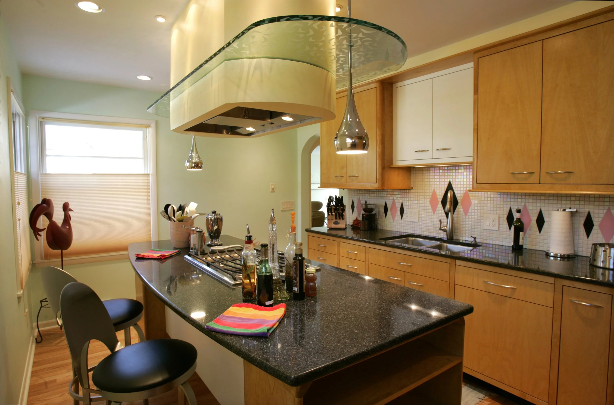 Modern kitchen with black granite island, light wood cabinets, and a decorative tile backsplash. There are two hanging pendant lights, a double sink, and various kitchen utensils and condiments on the counter. A nearby window has blinds, and a small 
