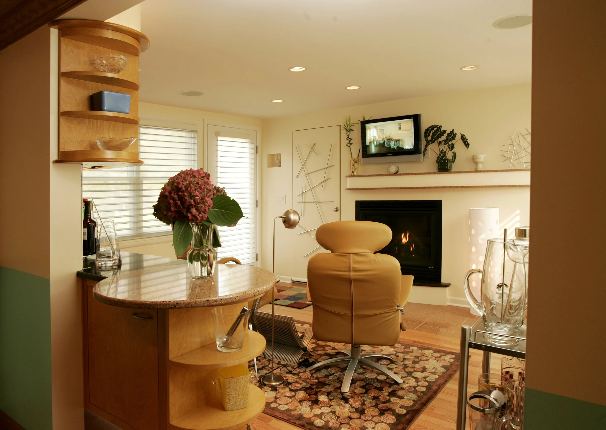Cozy living room with a fireplace, yellow armchair, and glass decor, viewed from the kitchen area with a vase of pink flowers on the counter.