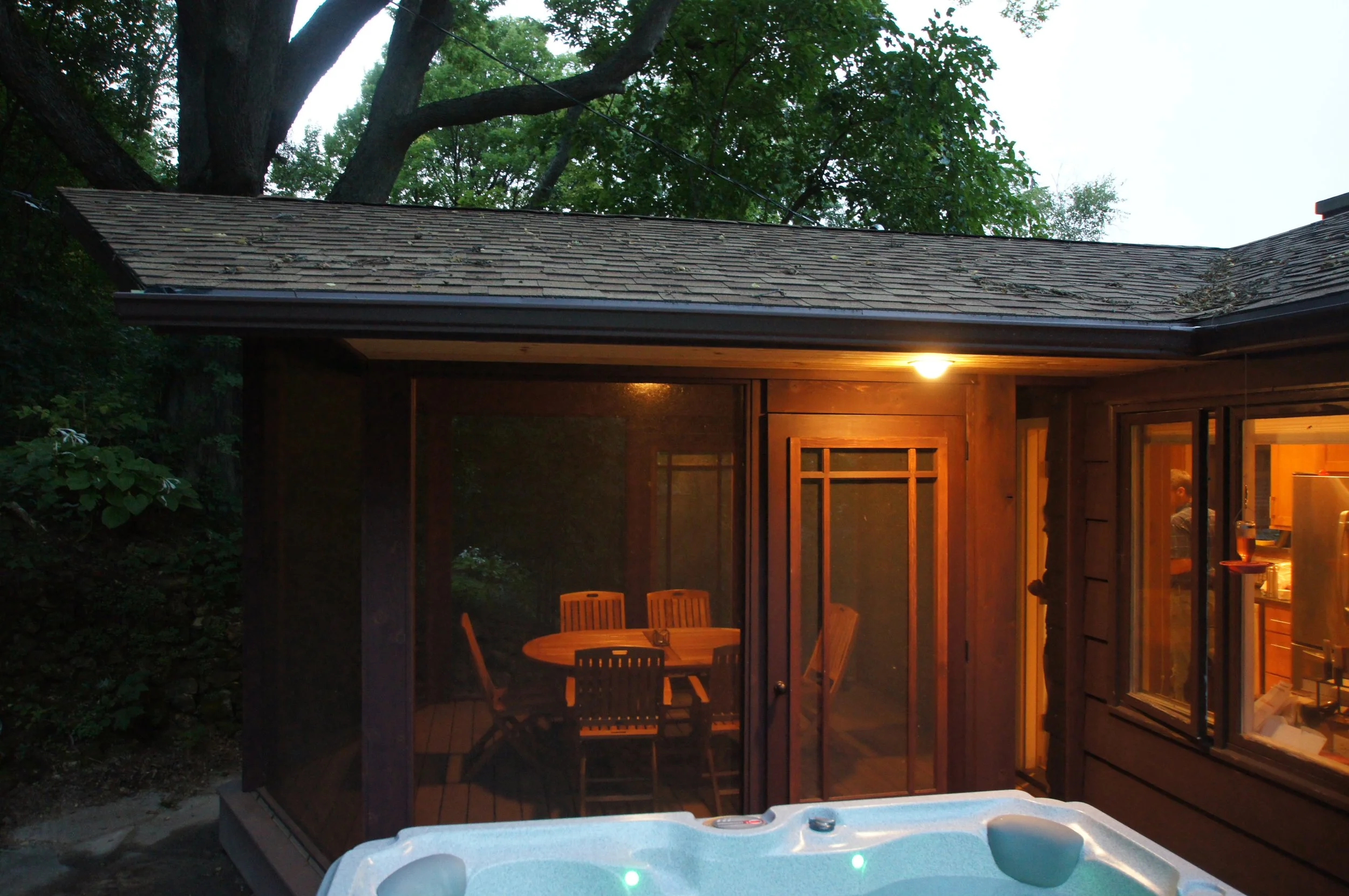 A backyard patio with a hot tub in the foreground and a screened porch attached to a house in the background, illuminated by warm indoor lighting at dusk or evening.