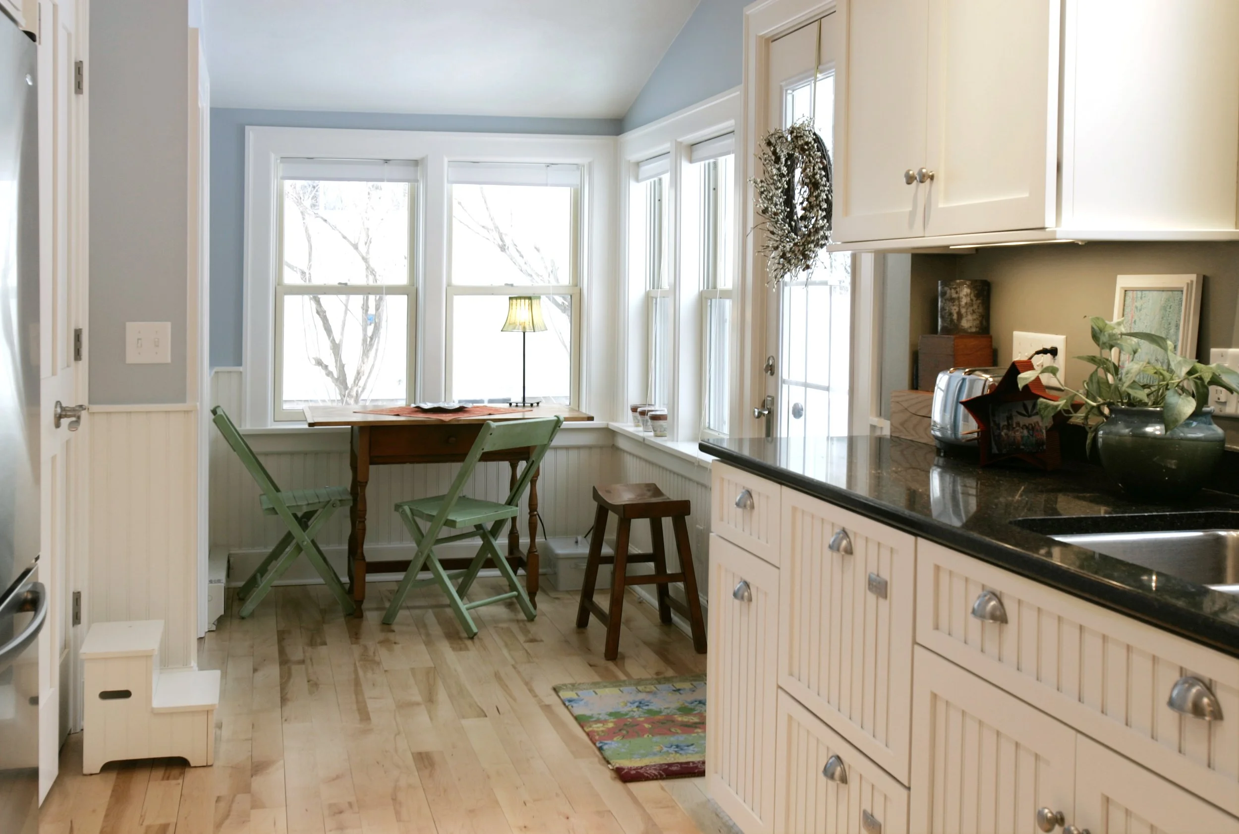 Bright kitchen with white cabinets, black granite countertops, and a small breakfast nook with a table, empty chairs, and a window seat. Decor includes a wreath on the door, plants, a lamp, and colorful rugs.