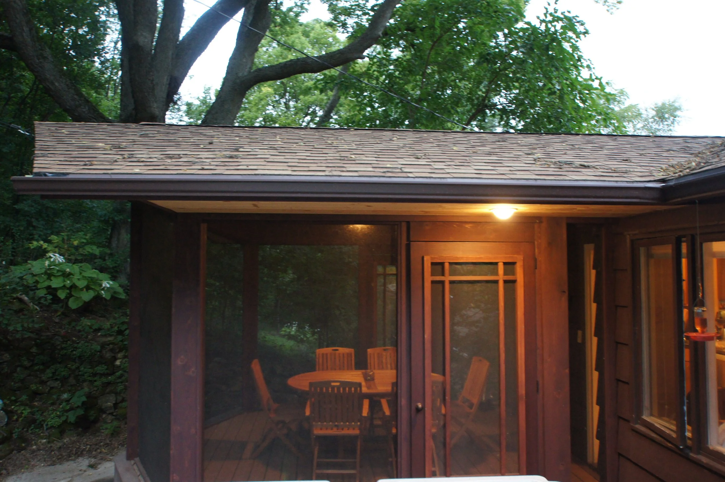 Back porch of a house with a screened-in area, wooden table, chairs, and an outdoor light, surrounded by trees and greenery.