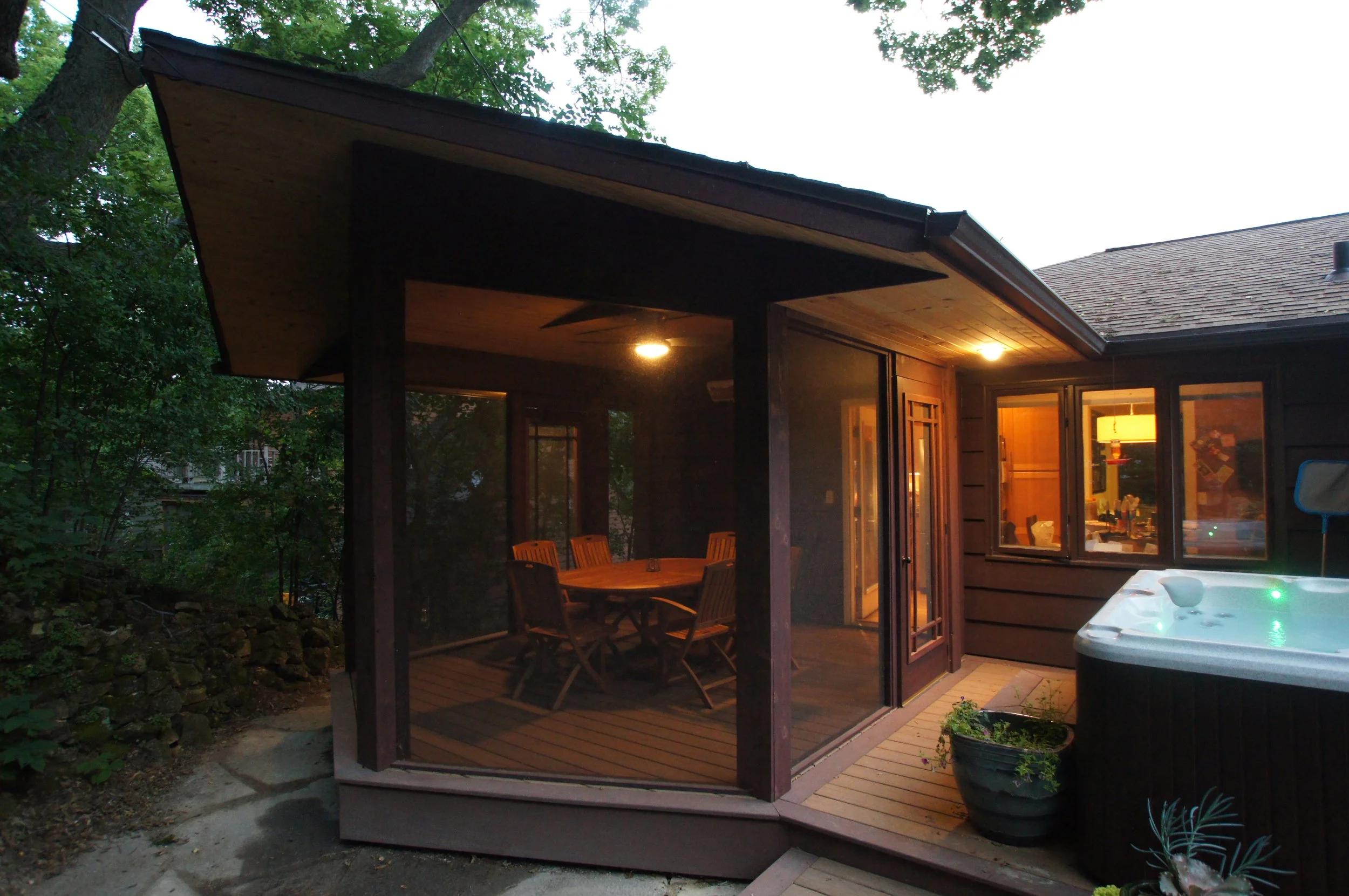A screened porch with a wooden dining table and six chairs, attached to a house with outdoor lighting, trees surrounding the area, and a hot tub on the deck.