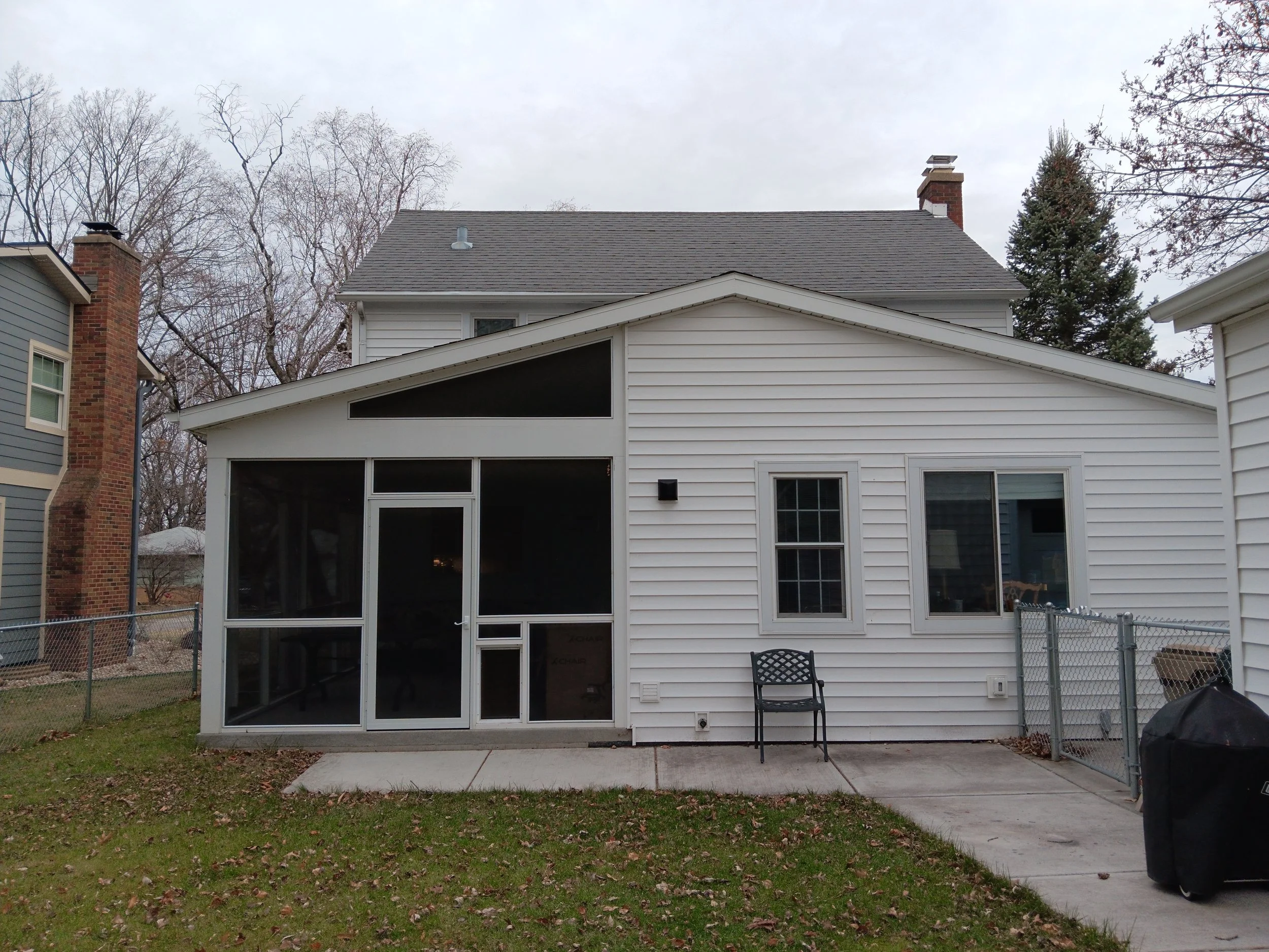 Rear view of a two-story house with white siding, featuring a screened-in porch with a sliding door, a small black chair outside, and a barbecue grill on the patio, surrounded by a lawn and neighboring houses.